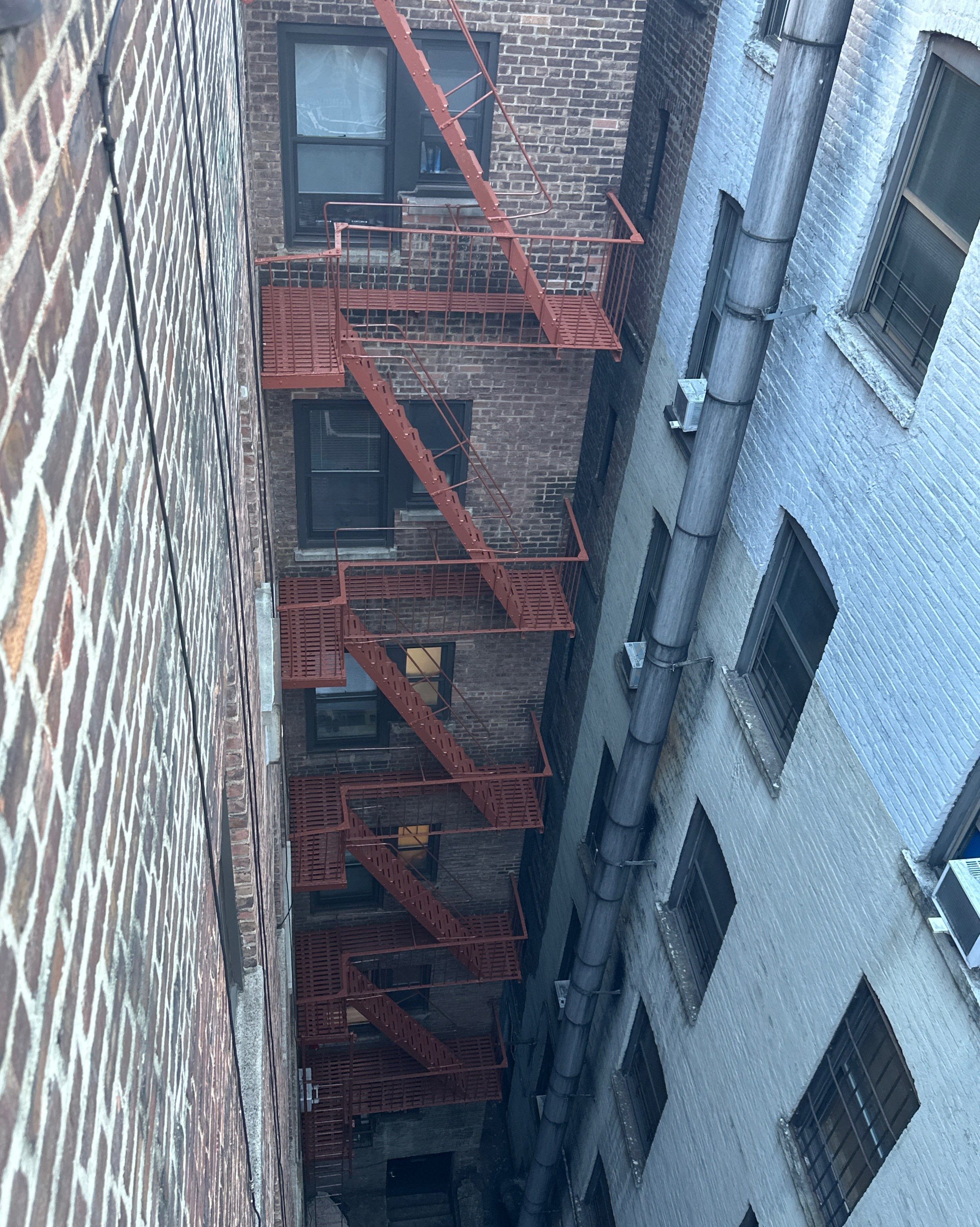 View of a fire escape with red stairs connecting four floors between two brick building walls in an alley.