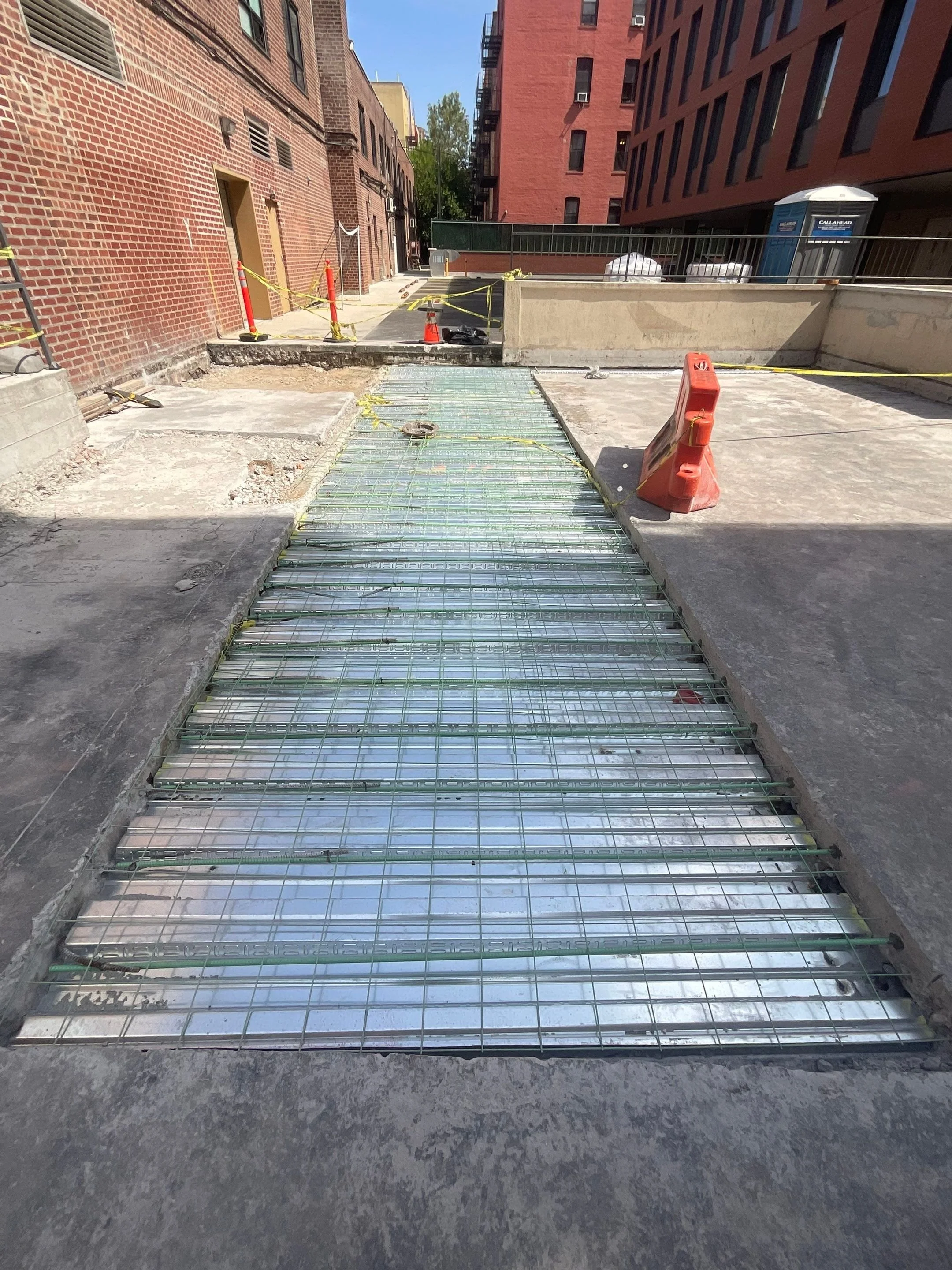 Construction site showing a section of exposed rebar and metal decking on a street, surrounded by safety cones, caution tape, and orange barricades, with brick and building structures in the background.