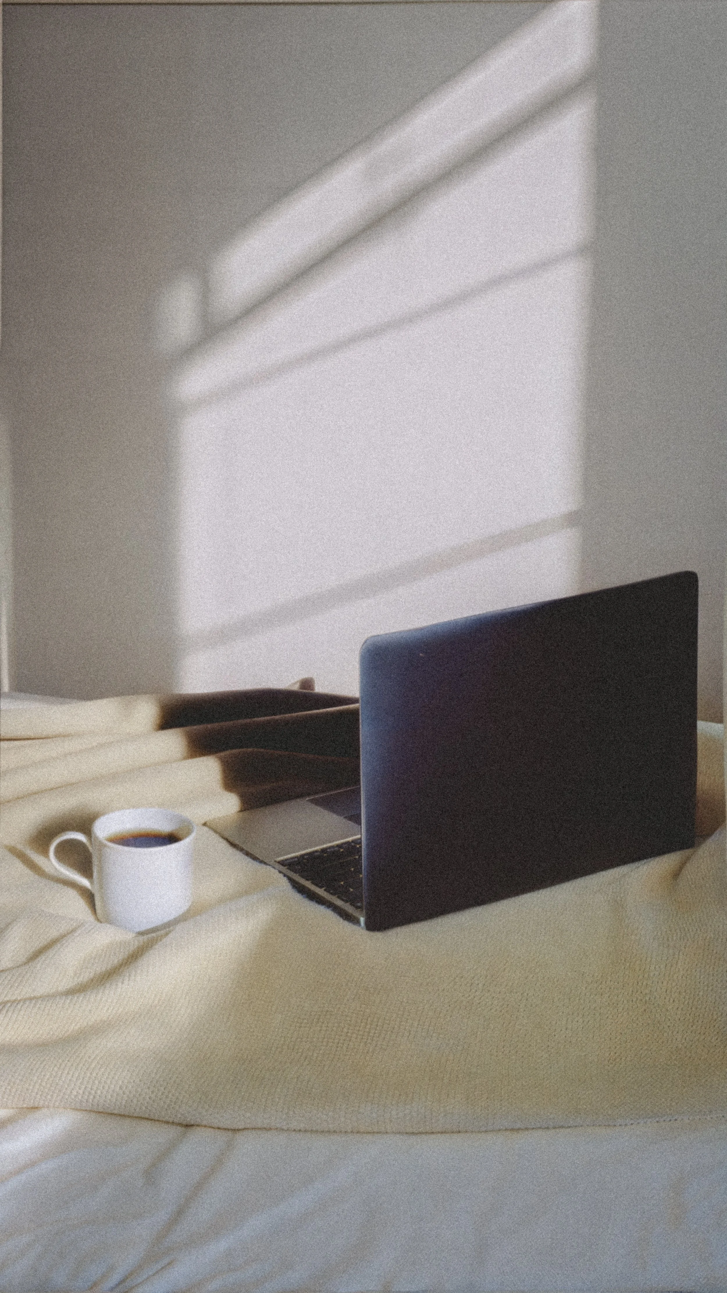 A laptop and a white coffee mug filled with coffee resting on a bed with beige bedding. Shadows cast from window blinds create horizontal lines on the wall.