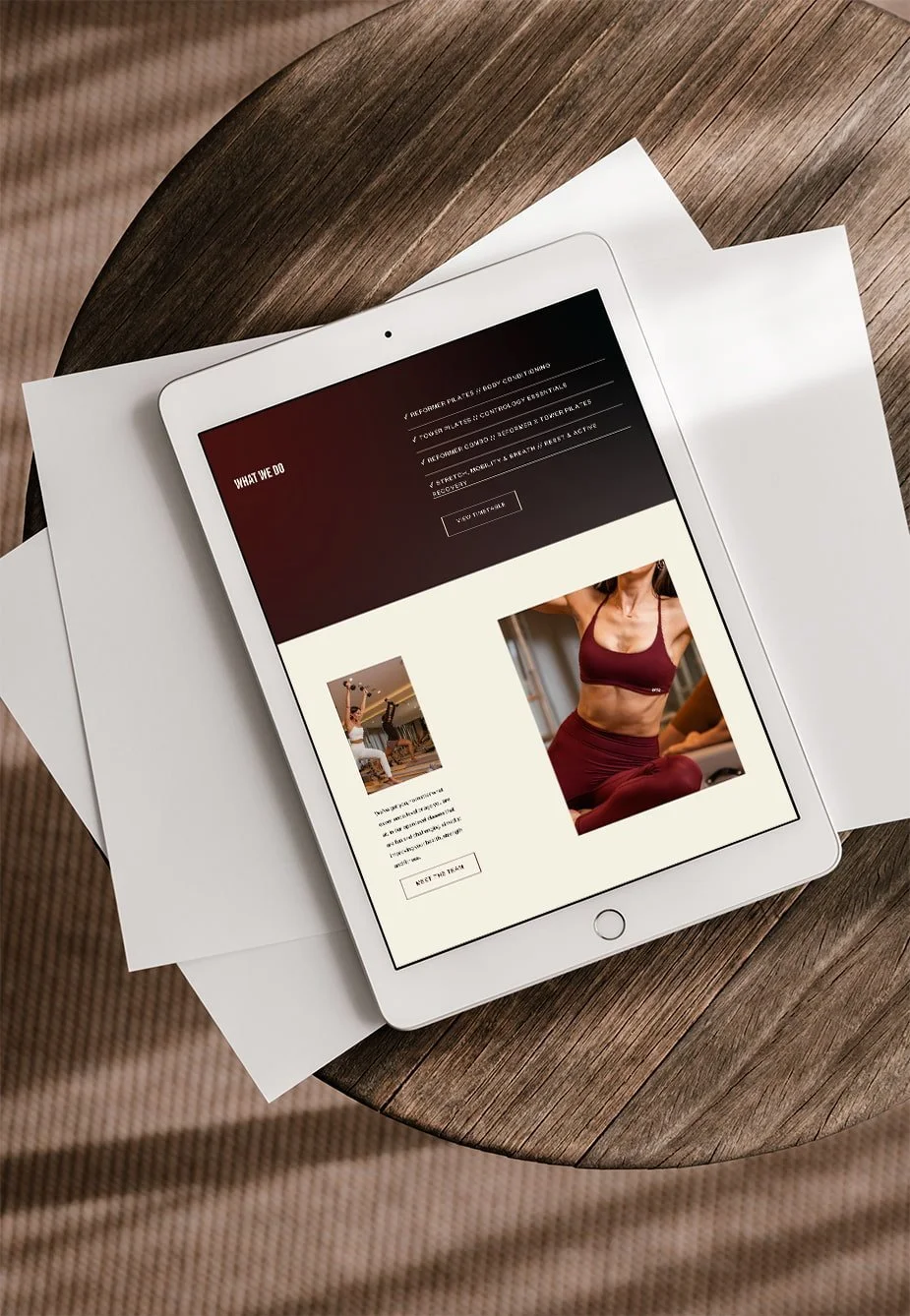 An iPad displaying a fitness website on a wooden table, with a paper underneath. The website features a woman in activewear and information about fitness programs.