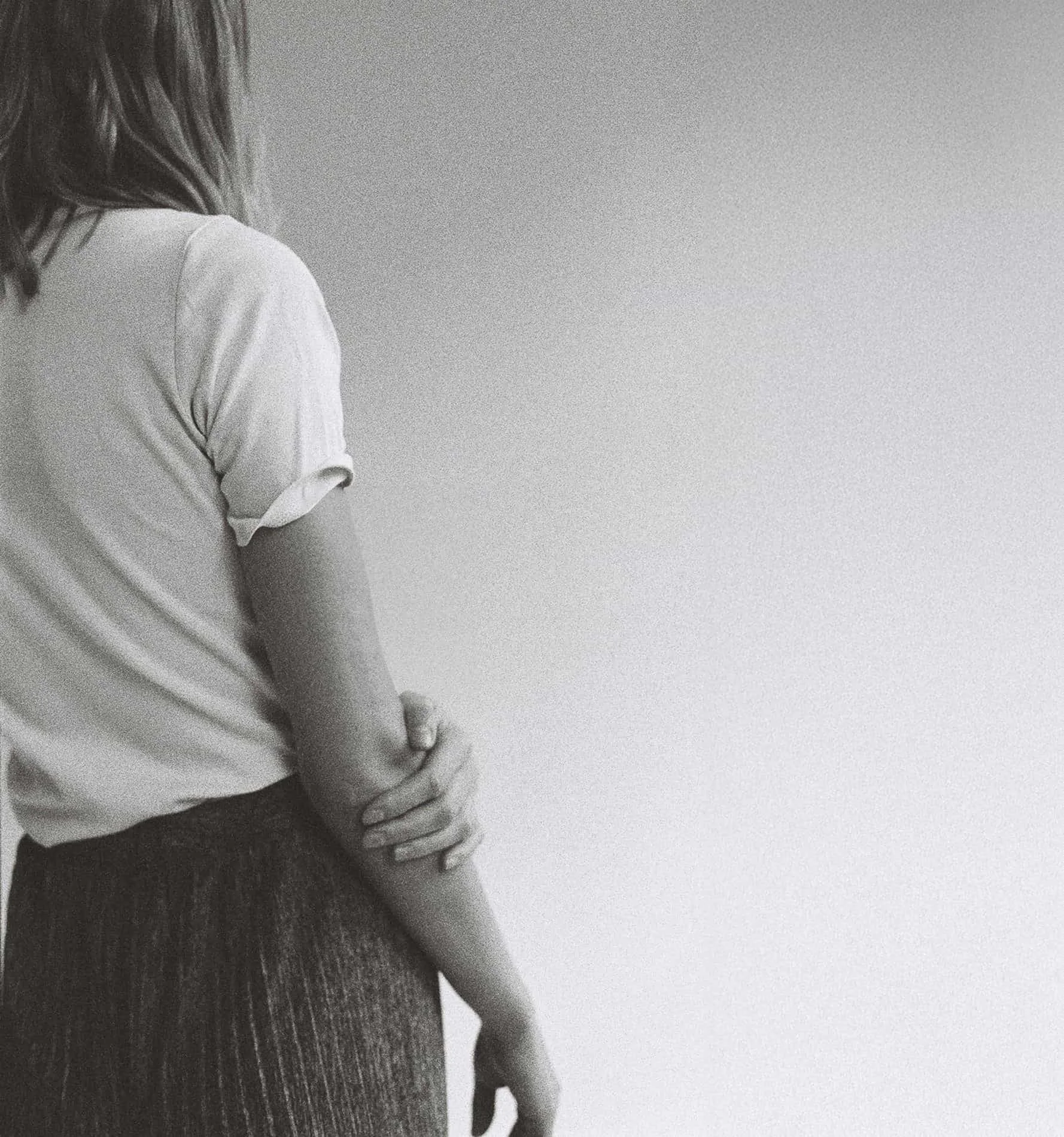 A person with shoulder-length hair wearing a light-colored T-shirt and a dark striped skirt, holding their left elbow with their right hand, standing against a plain light background.