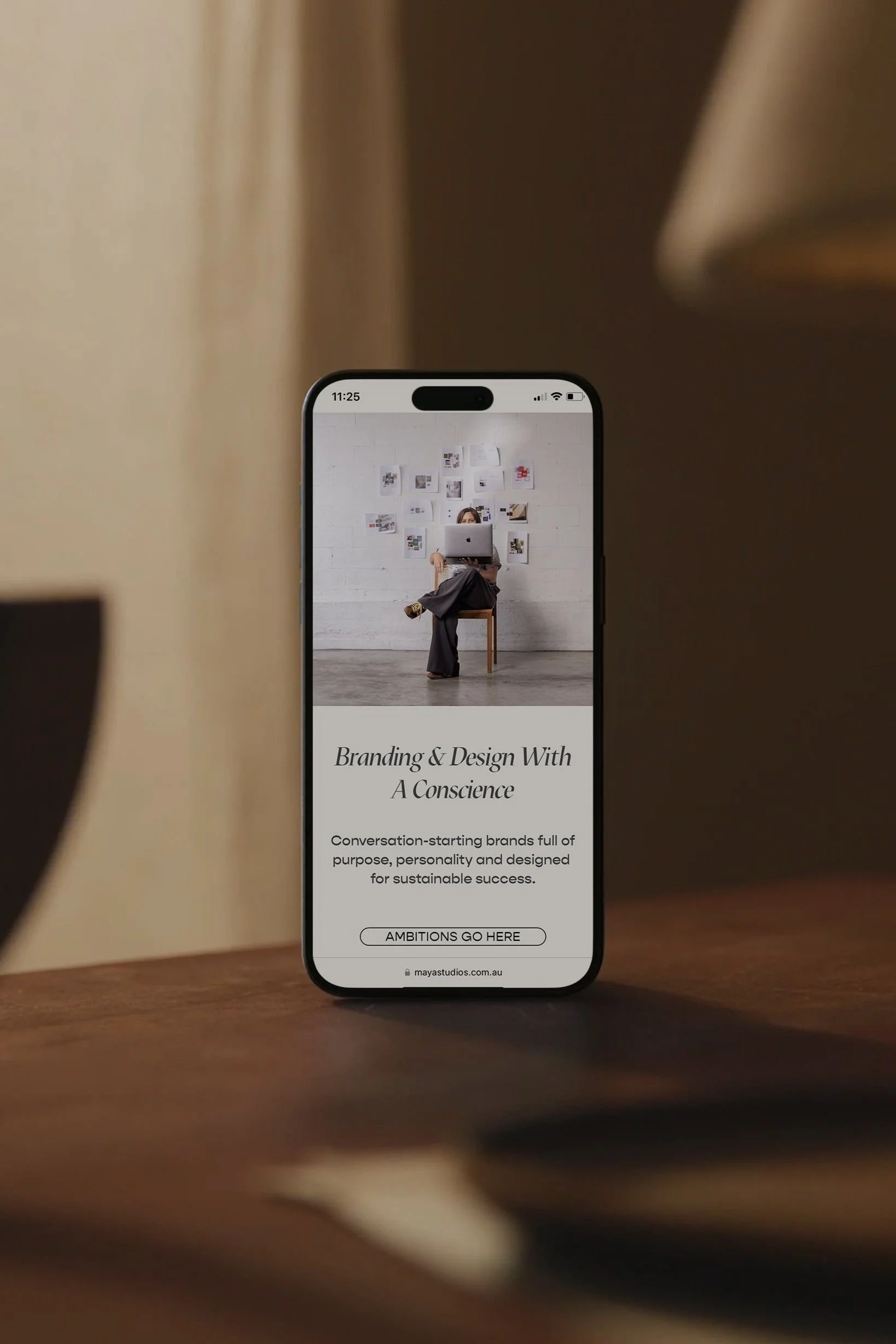 A smartphone displays a webpage titled 'Branding & Design With A Conscience,' showing a woman sitting on a chair with a laptop, and a white wall with pictures behind her. The webpage includes a button labeled 'AMBITION GO HERE' and a website URL 'mayastudios.com.au'.