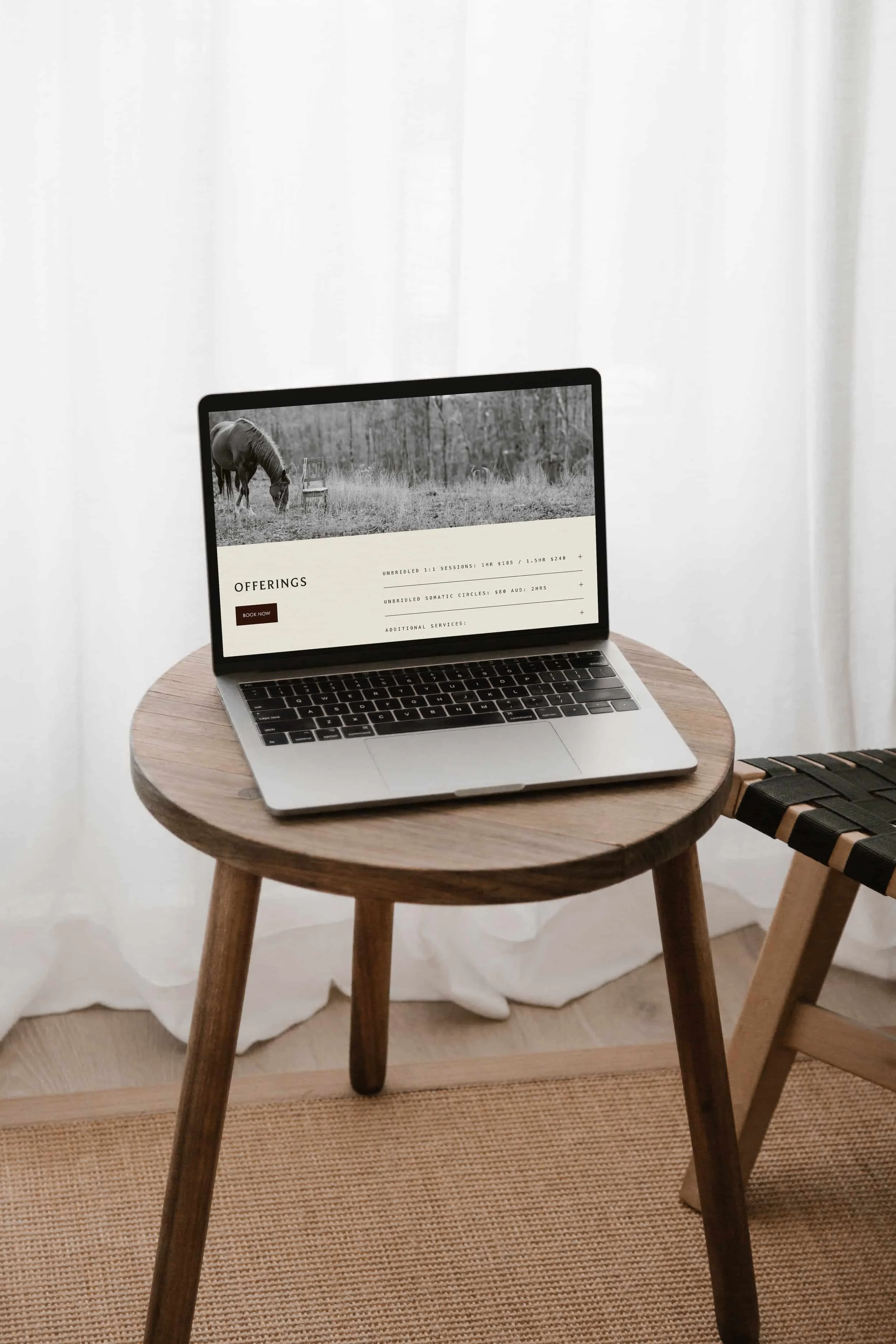 A laptop on a wooden table displaying a website with offering details, next to a woven chair and white curtains in a bright room.