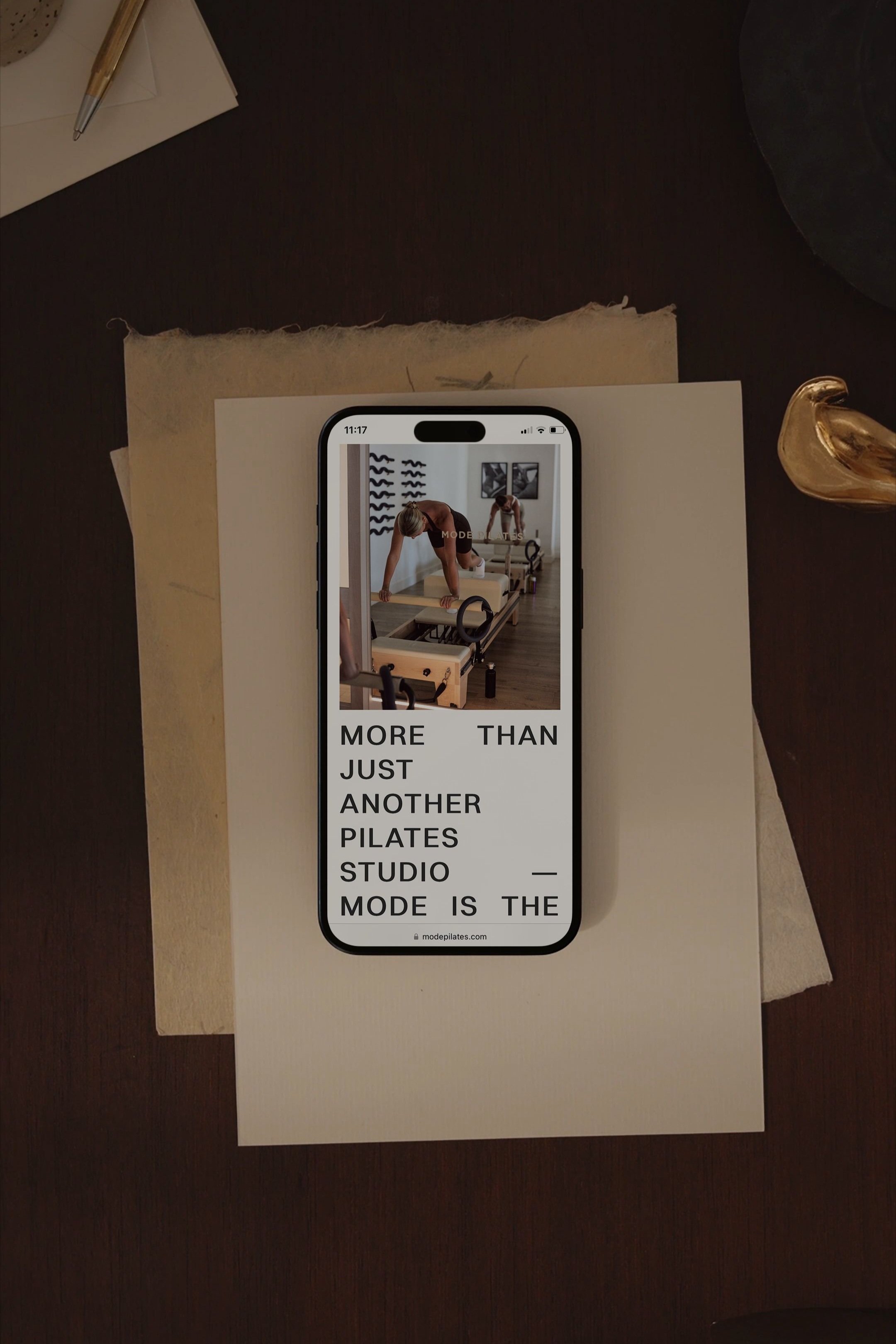 A smartphone on a desk displaying a Pilates studio with two instructors, and overlay text reading "More than just another Pilates studio — mode is the".