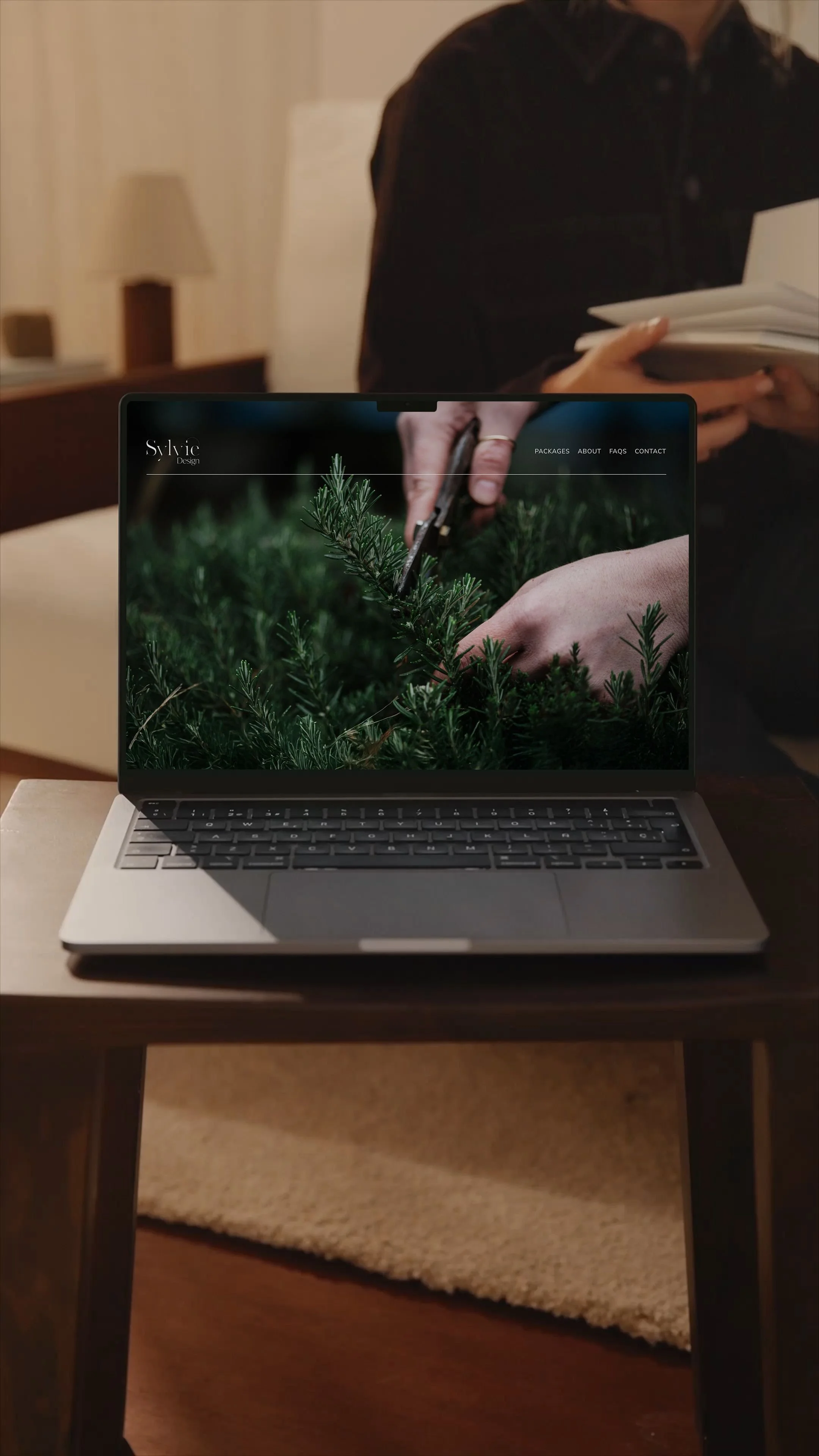 Laptop displaying a website with an image of hands cutting a Christmas tree branch, placed on a wooden table. Behind, a person holds a notebook in a cozy indoor setting.