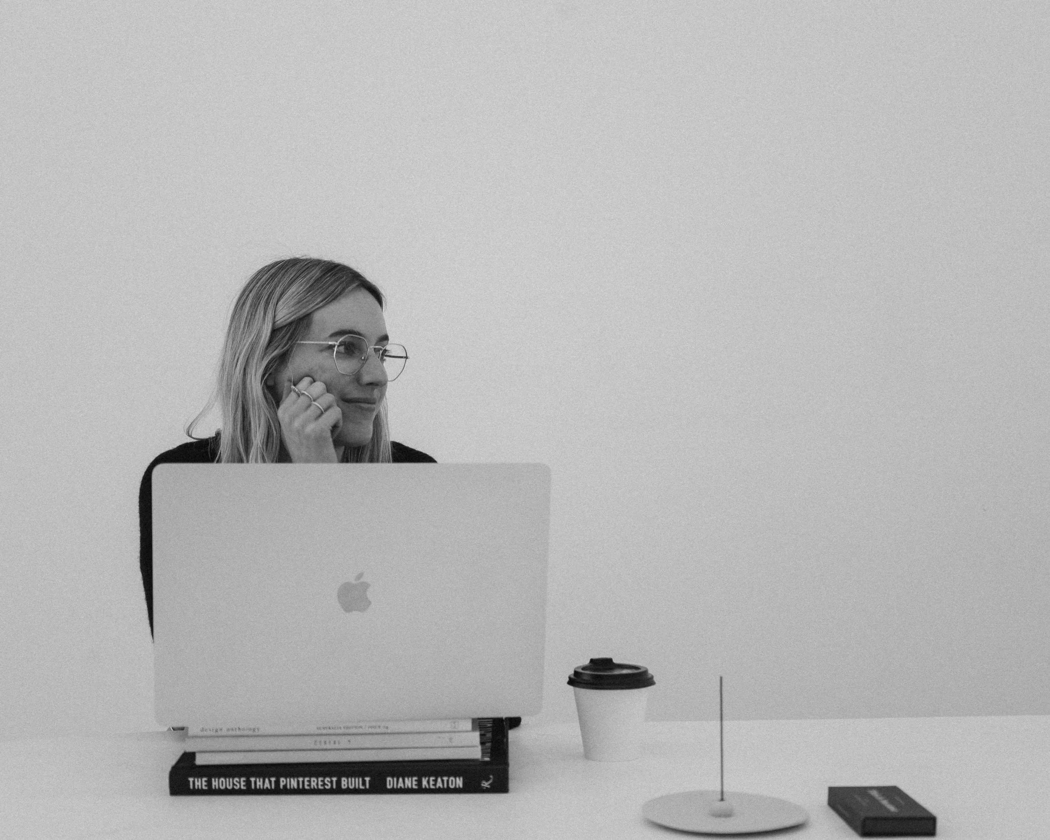 A woman with glasses sitting at a desk with a laptop, a cup, and two books, one titled 'The House That Pinterest Built' by Diane Keaton