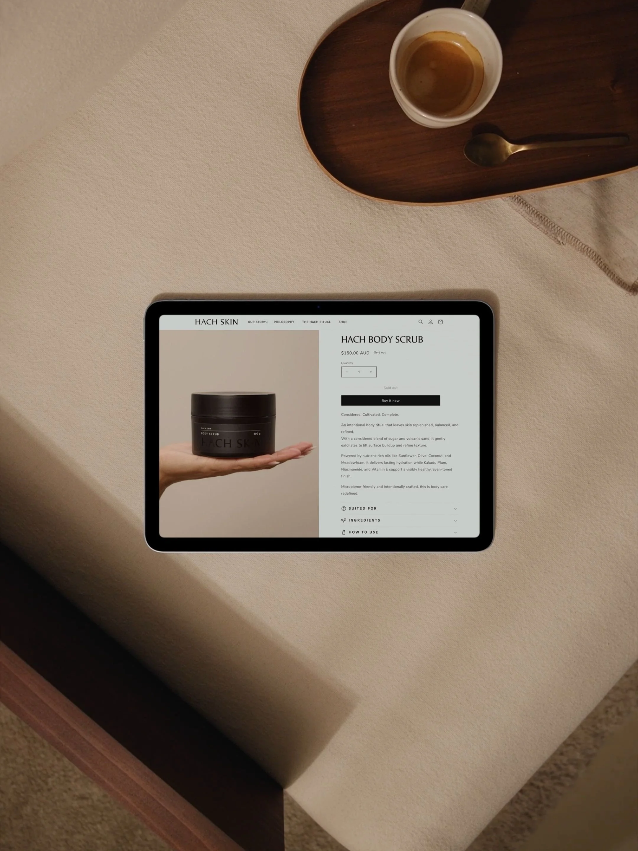 An iPad displaying a webpage for Hach Skin Body Scrub on a beige table. A white cup with an empty espresso shot and a small gold spoon on a wooden tray are nearby.