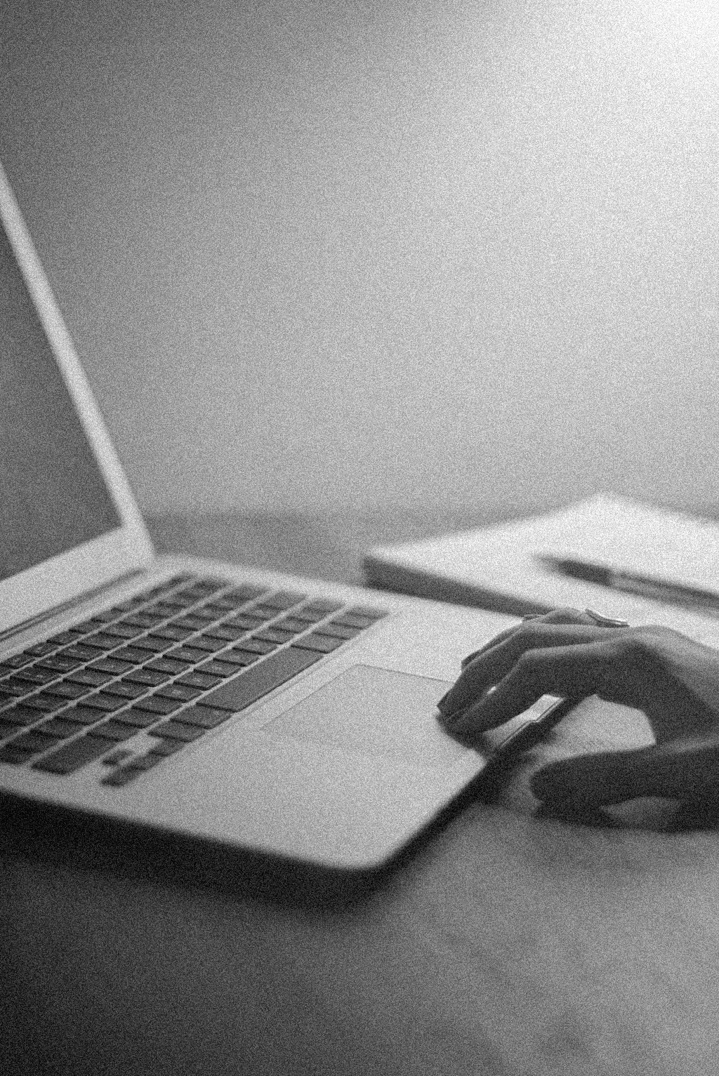 Black and white photo of a person's hand on a laptop keyboard, with a notebook and pen on the desk nearby.