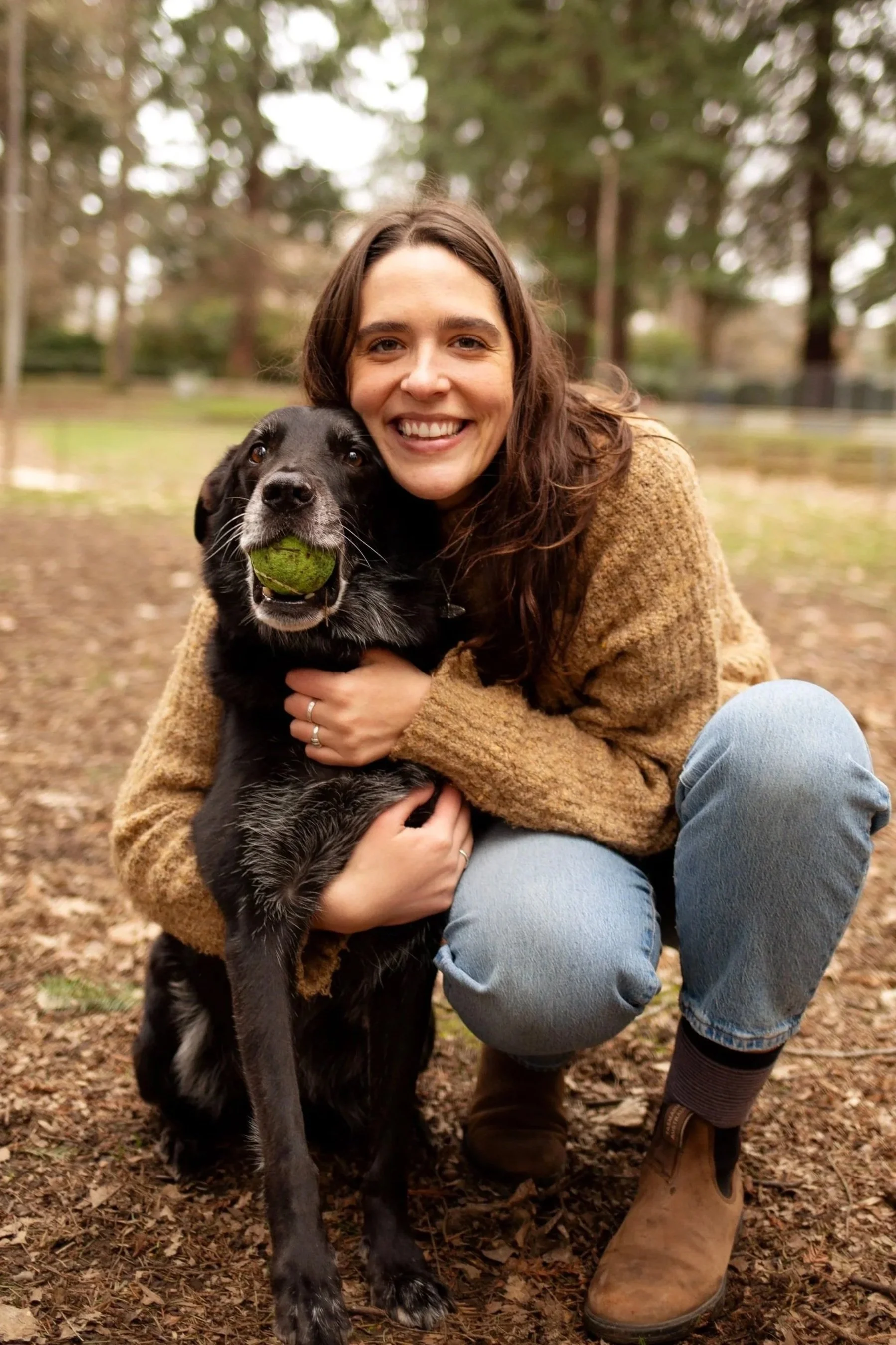 A woman smiling outdoors squatting with a black dog holding a tennis ball in its mouth, on a background of trees and autumn ground.