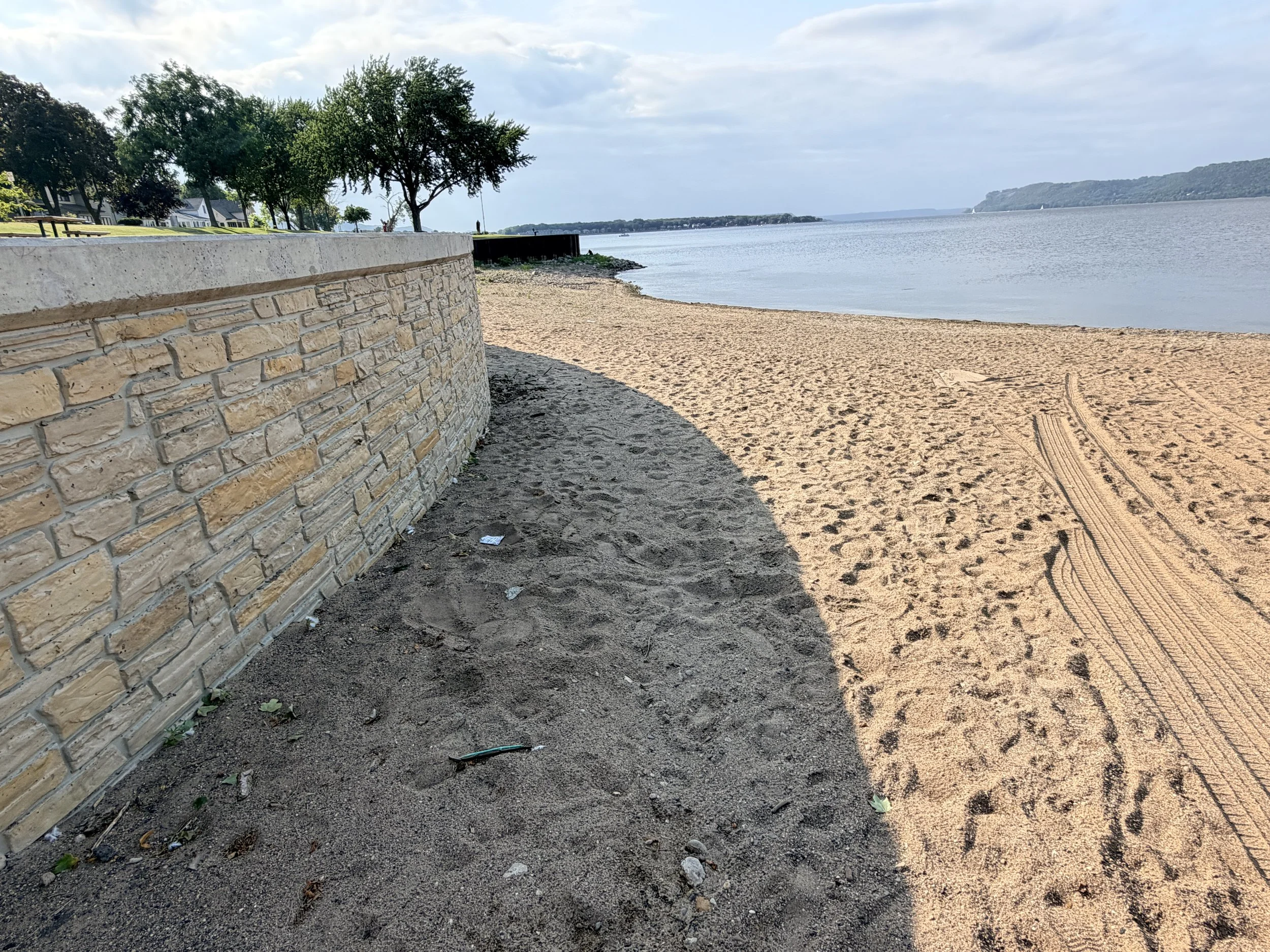 View of a beach with sandy shore, a brick stone wall on the left, and water extending into the horizon with distant land and a cloudy sky.