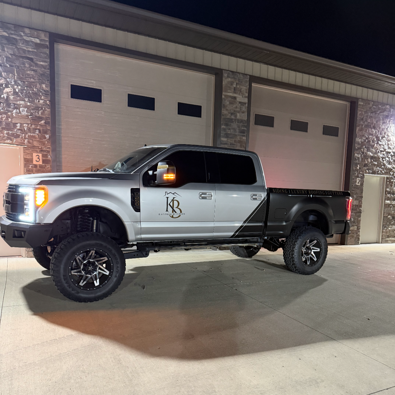 A lifted pickup truck parked outside a building at night, with a black and white color scheme, custom wheels, and a logo on the door that reads 'KB' with the words 'Kaczmarz Builders' beneath.