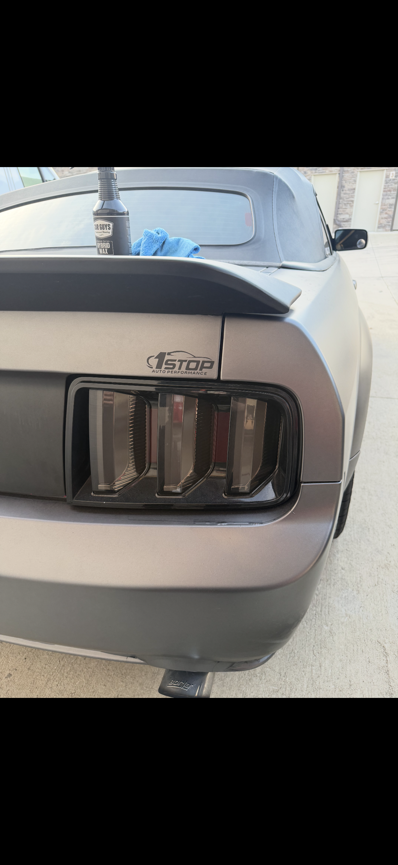 Rear view of a silver Ford Mustang with black taillights and a 1 STOP auto performance badge, with cleaning supplies on the trunk, including a spray bottle and a blue cloth.