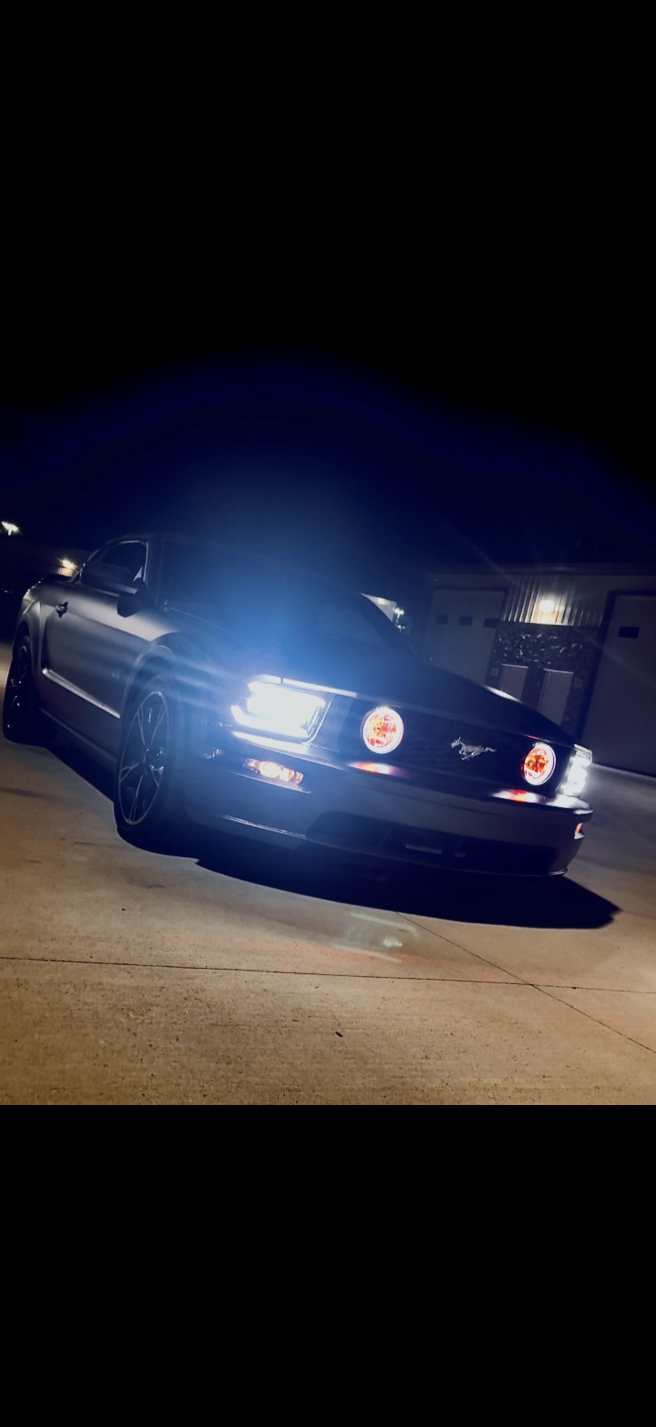 A black Ford Mustang parked on a driveway at night, illuminated by its headlights, with a house and garage in the background.