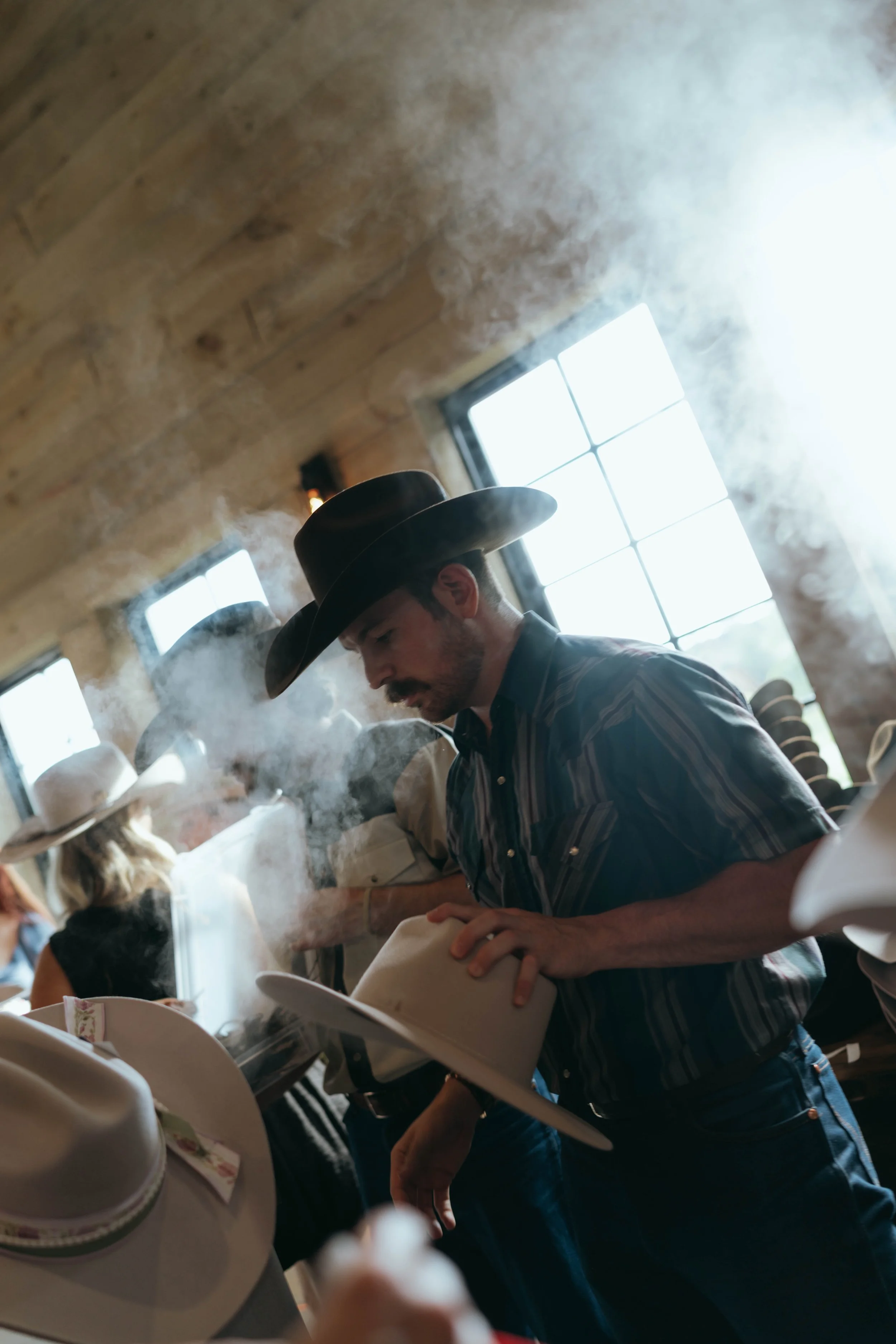 A man wearing a cowboy hat shapes a felt fedora hat with steam.