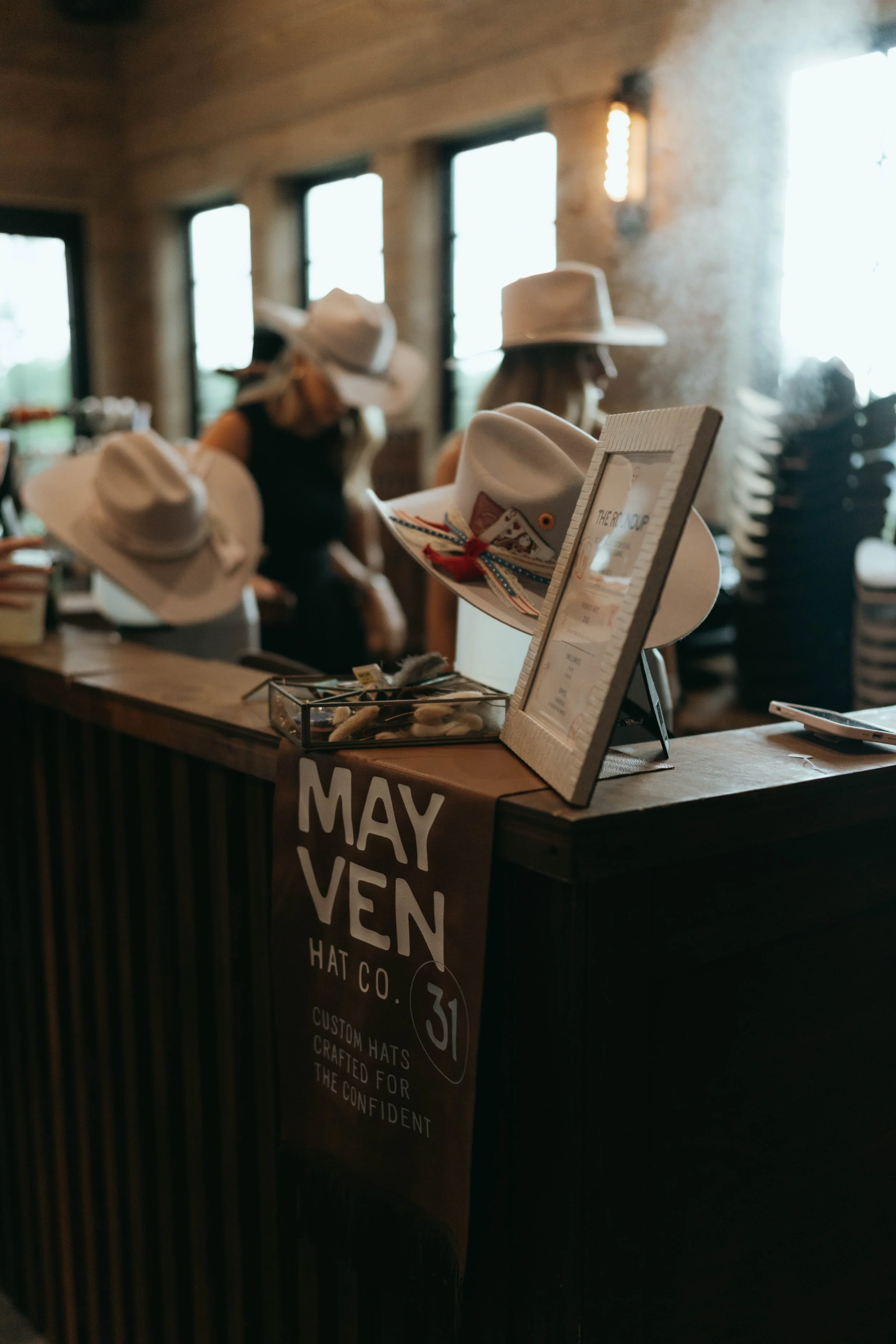 A wooden bar set up with felt cattleman and fedora hats with two women behind the bar creating custom hats.