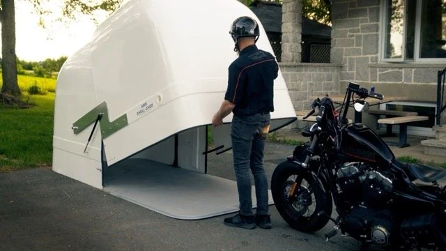 Une personne en casque regarde une structure blanche, probablement une unité de désactivation de drone ou un abri technologique, avec une moto noire garée à côté.