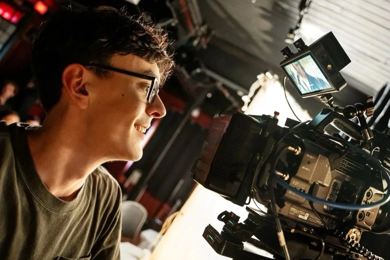 Un homme portant des lunettes, souriant, assis devant une caméra professionnelle dans un studio, en train de filmer ou de préparer une prise.