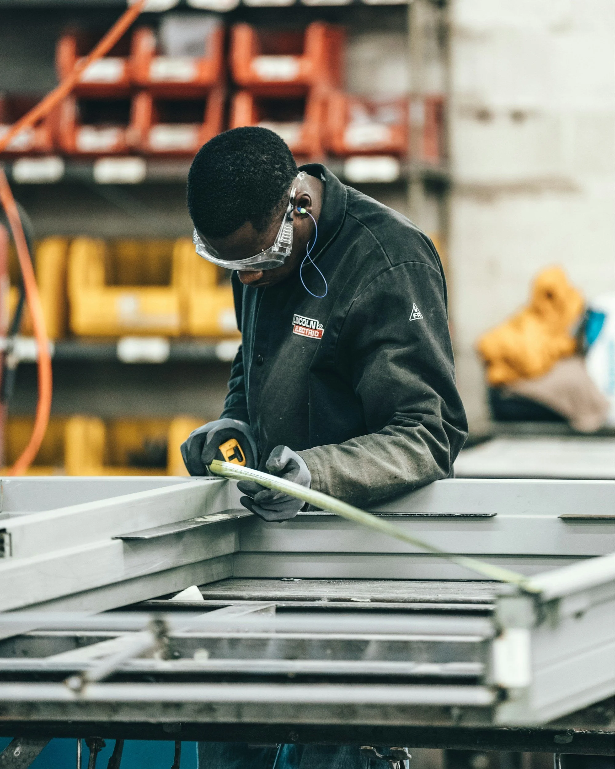 A worker in safety goggles and gloves measuring a metal frame in a workshop or factory setting.