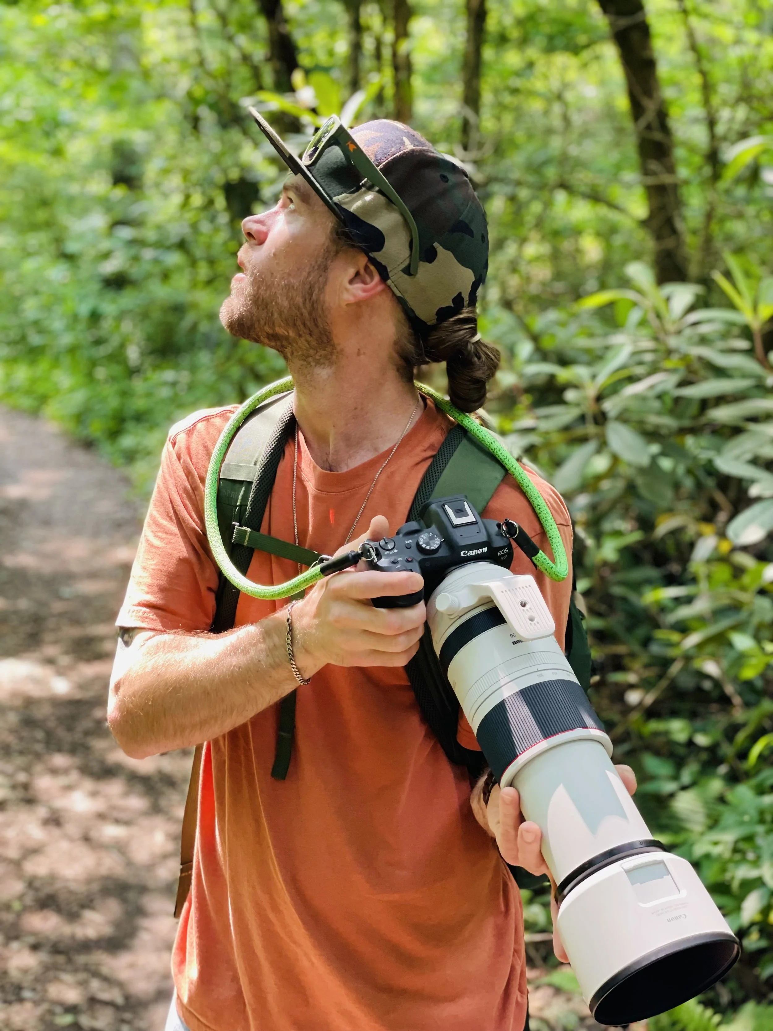 Man with a camera and a green hose wrapped around his neck, standing on a wooded trail, looking upwards.