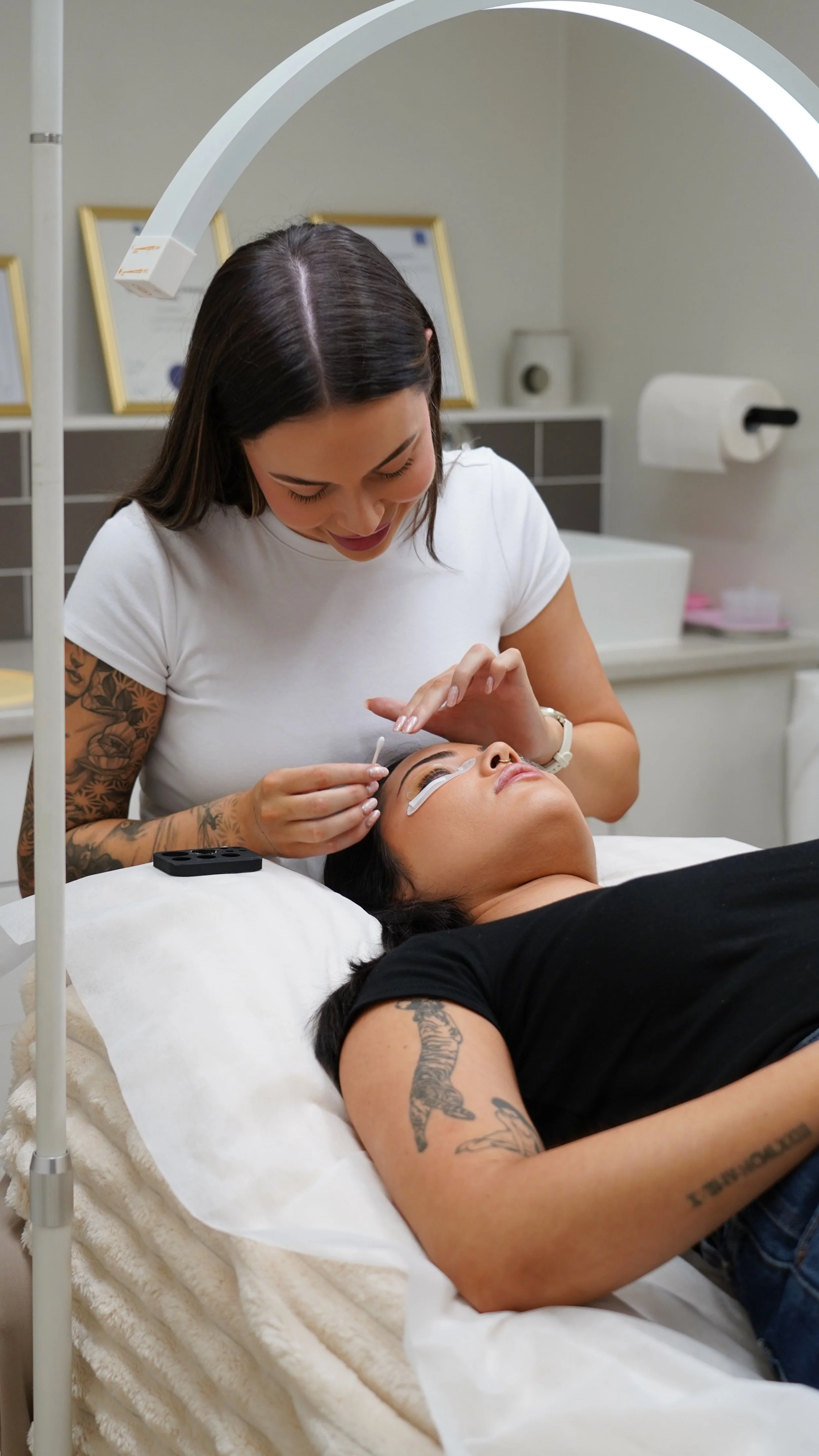 A woman sitting on a modern white chair with a rattan backrest, barefoot, using a pink Microsoft laptop, smiling, in a minimalistic room with light wooden floors and a white wall.