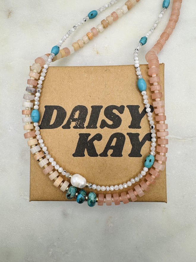 Two beaded necklaces, one with round pink and white beads and the other with turquoise and white beads, draped over a small cardboard box labeled 'Daisy Kay'.