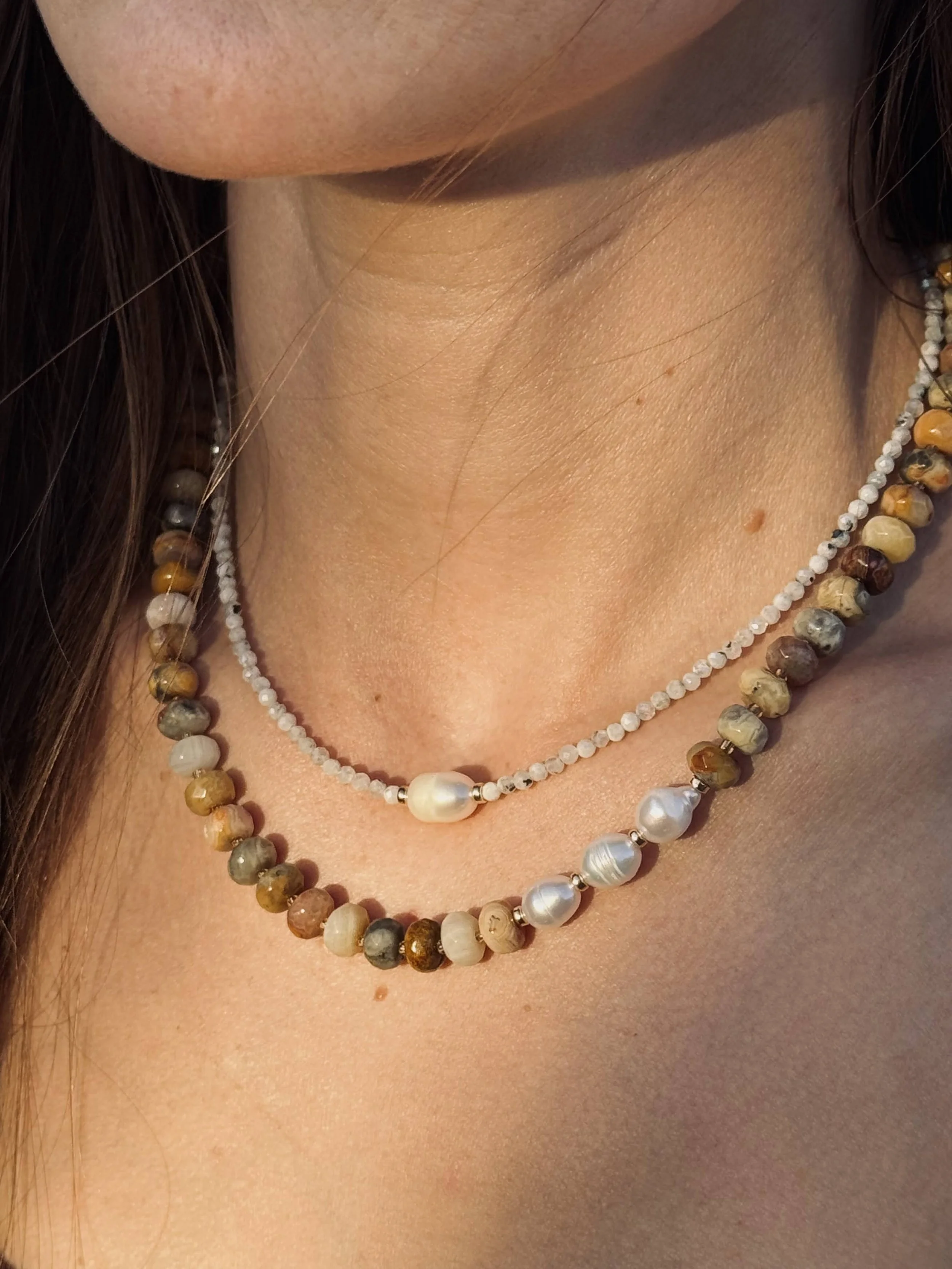 Close-up of a woman's neck with two beaded necklaces, one with small white beads and a large pearl, and the other with multicolored round beads and pearls.