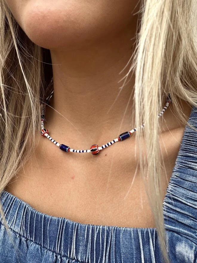 Close-up of a woman's neck wearing a beaded red, white, and blue necklace, with part of her blonde hair and a blue ruffled top visible.