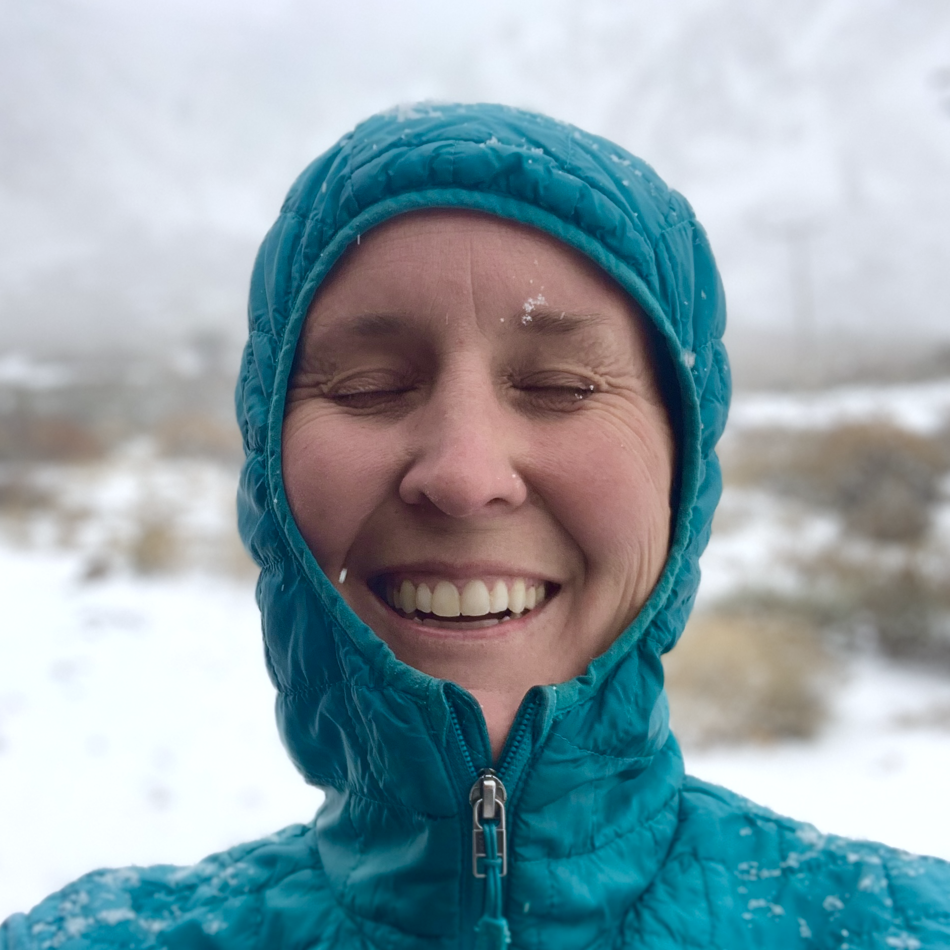 Close-up of a person smiling with eyes closed, wearing a bright blue winter jacket and matching hood on a snowy day.