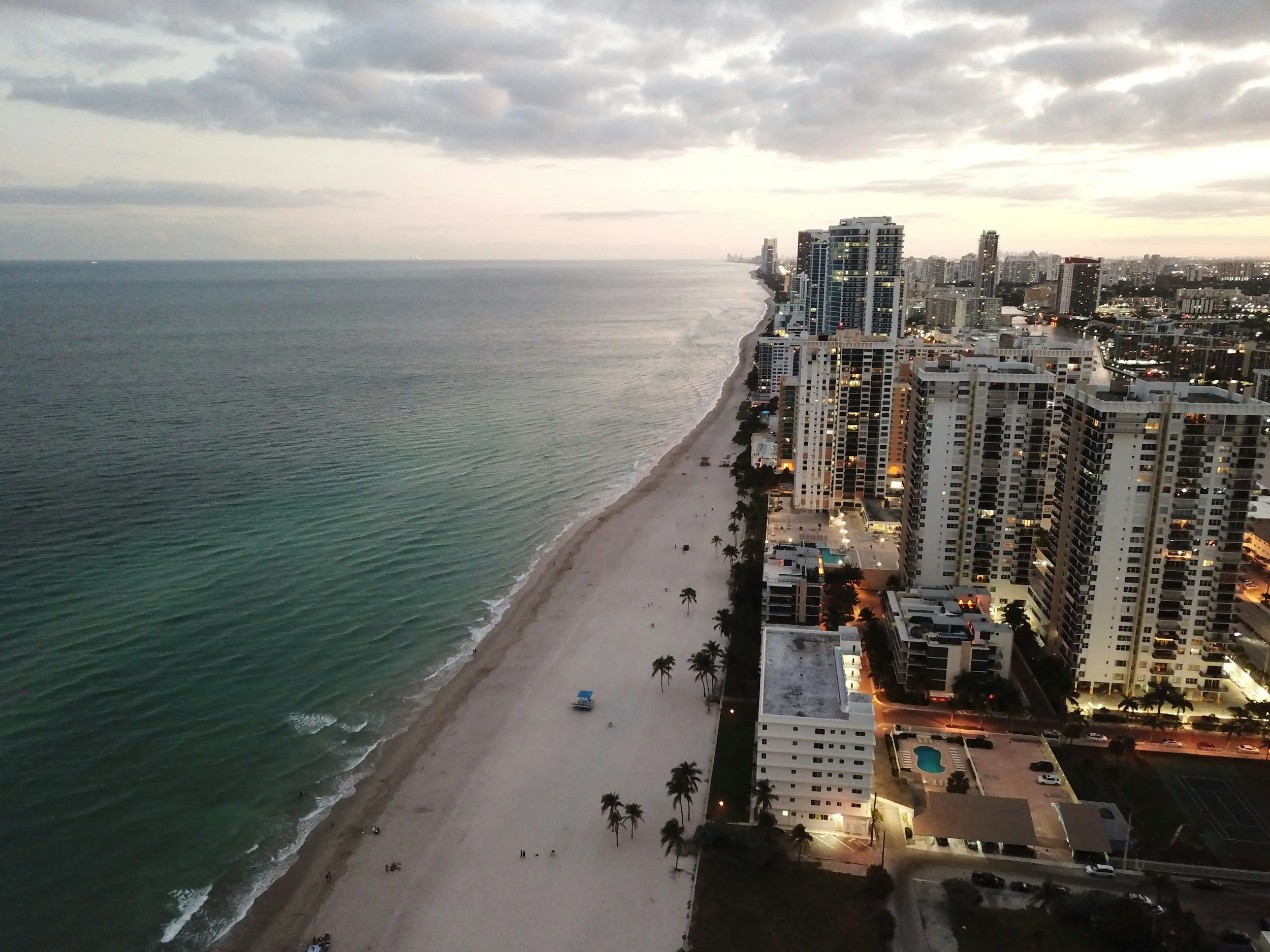 An aerial view of a city beachfront at sunset, showing tall skyscrapers, a sandy beach with a few palm trees, and the calm ocean waves stretching into the horizon.