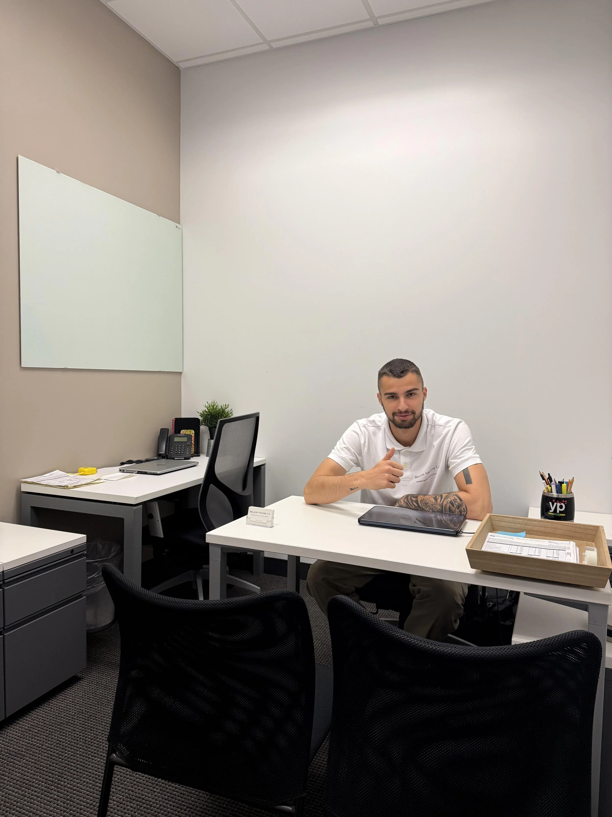 A man with tattoos on his right arm, sitting at a white office desk, giving a thumbs-up gesture, in a modern office.