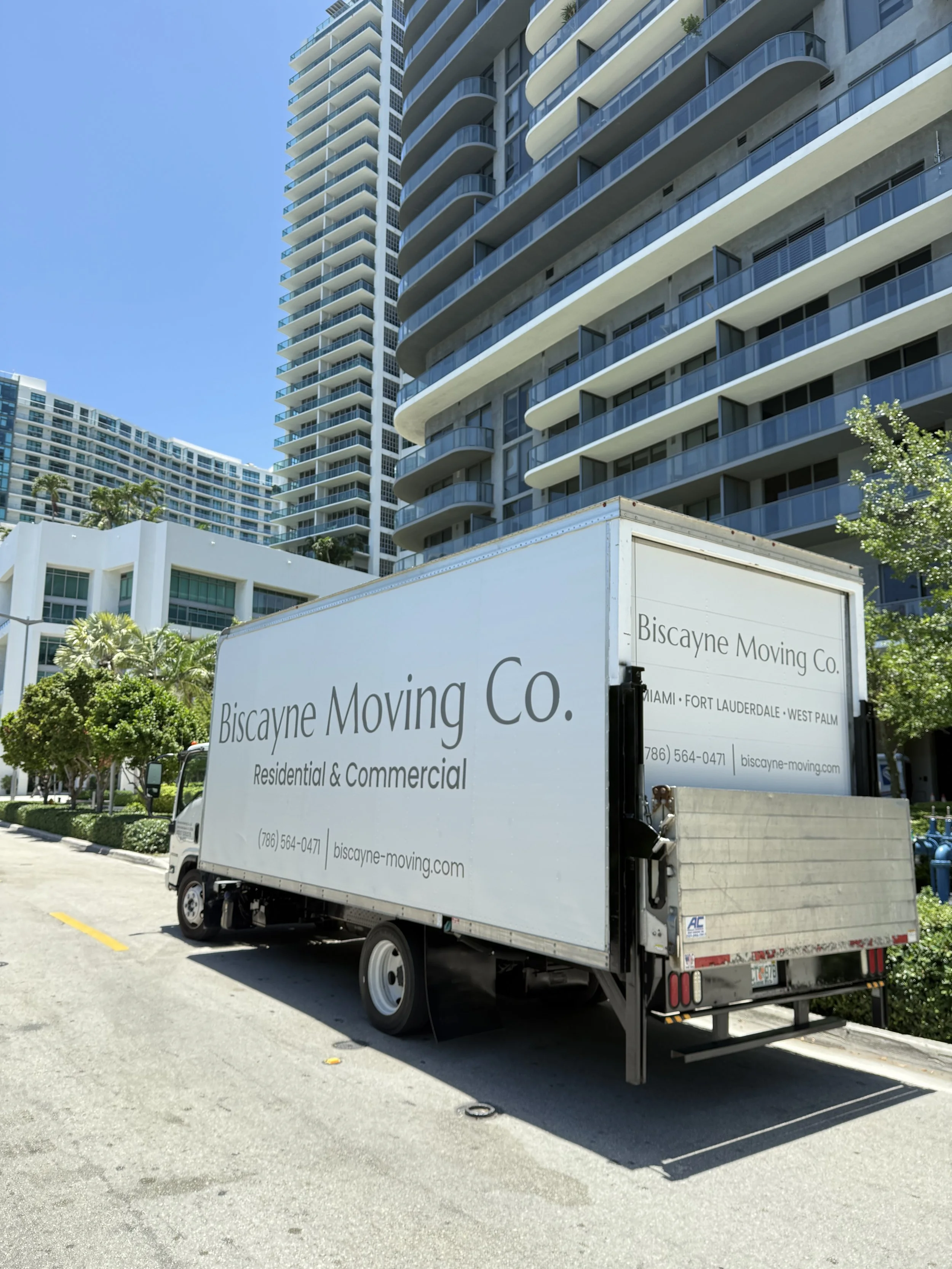 Moving truck with Biscayne Moving Co. logo parked in front of modern high-rise buildings with balconies, blue sky, and trees in Miami.