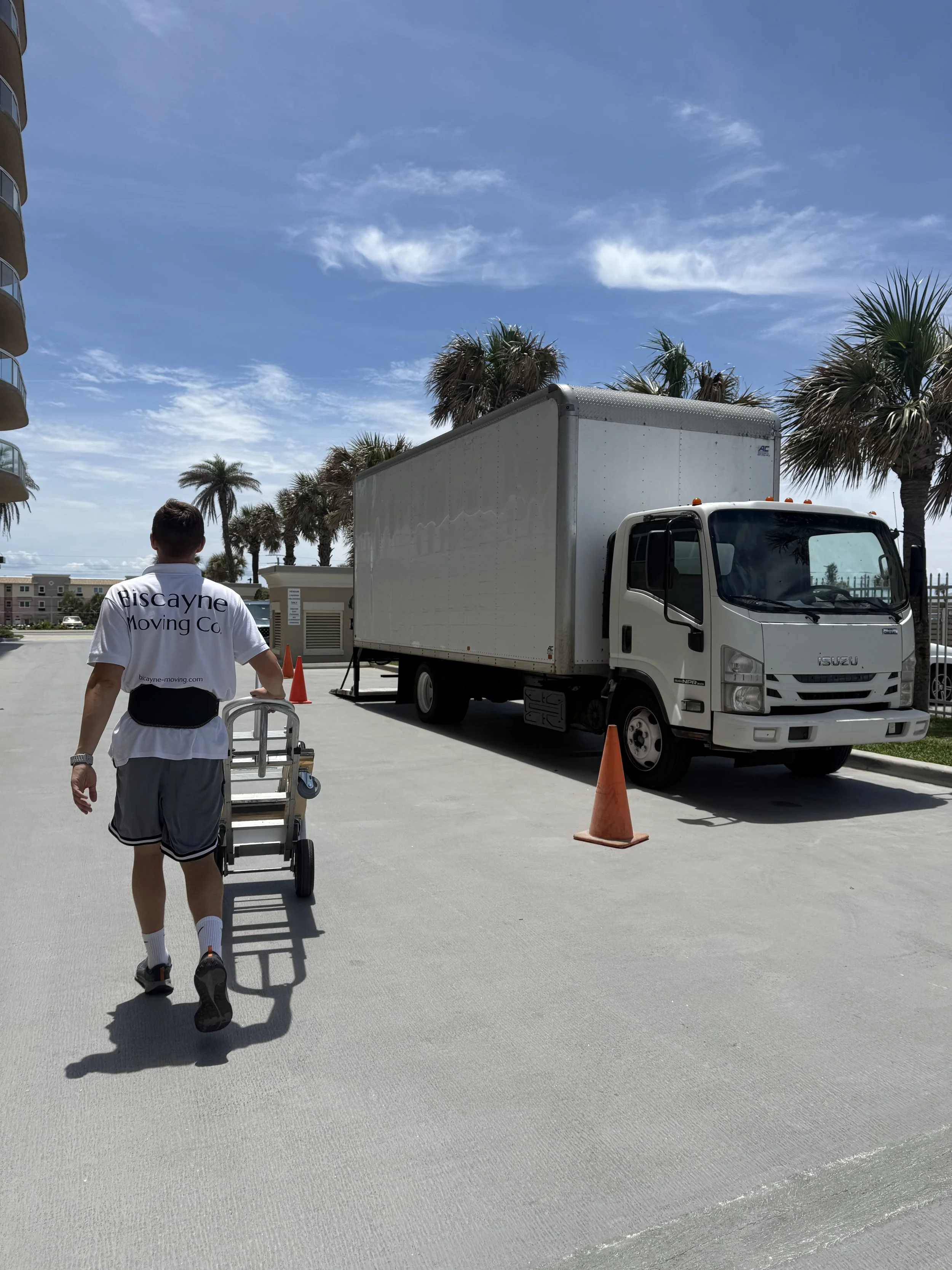 A man in a white shirt with 'Biscayne Moving Co.' printed on it, pushing a hand truck on a concrete pavement near a white moving truck and orange traffic cones, with palm trees and a blue sky with some clouds in the background.