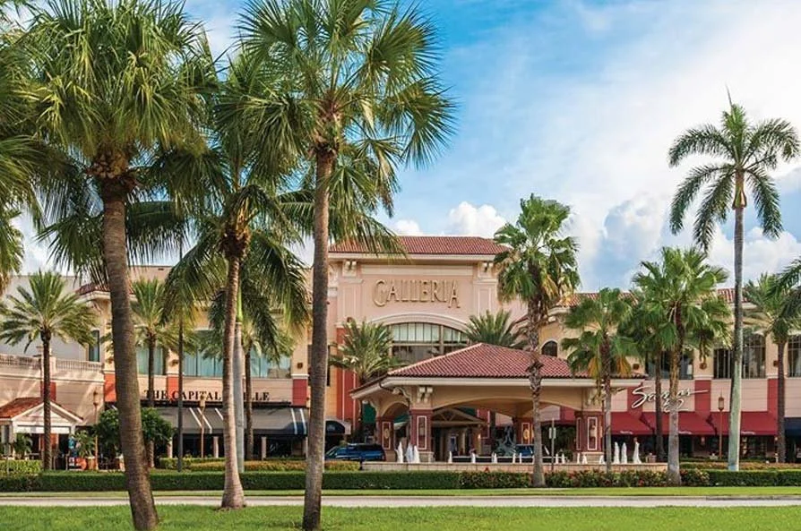 View of a shopping mall with palm trees in front, labeled 'GALLERIA,' with storefronts and a blue sky.