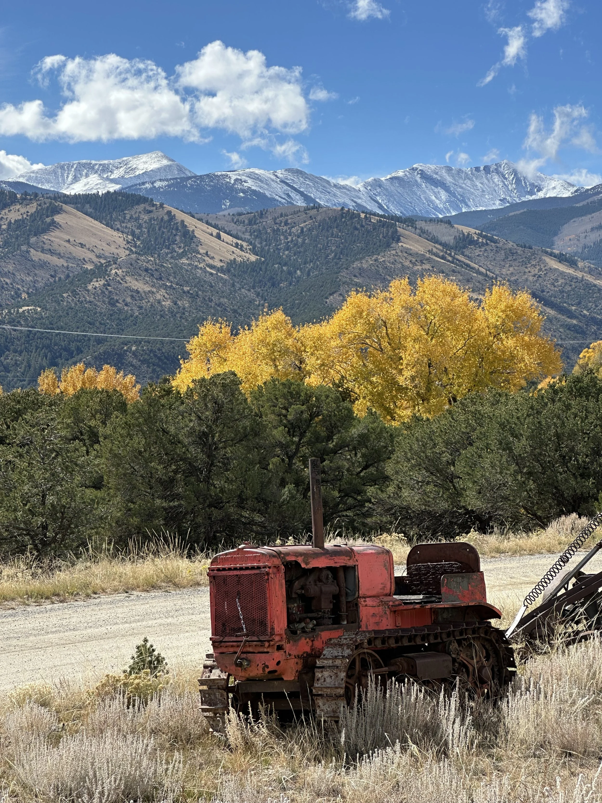 Old rusty red tractor sitting in dry grass with colorful trees and mountains in the background, snow-capped peaks under a blue sky with white clouds.