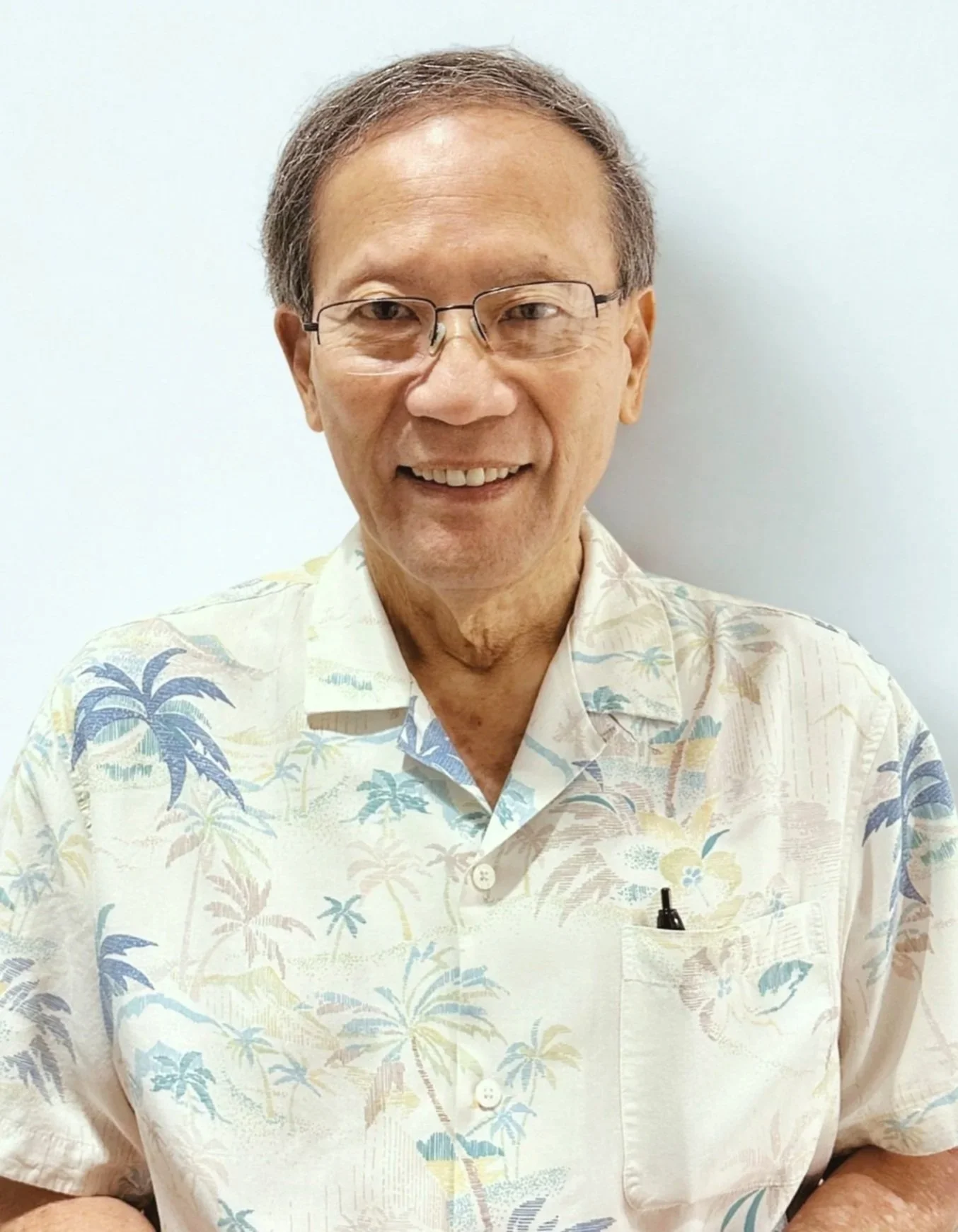 A smiling older man wearing glasses and a floral-patterned Hawaiian shirt standing against a plain white wall.