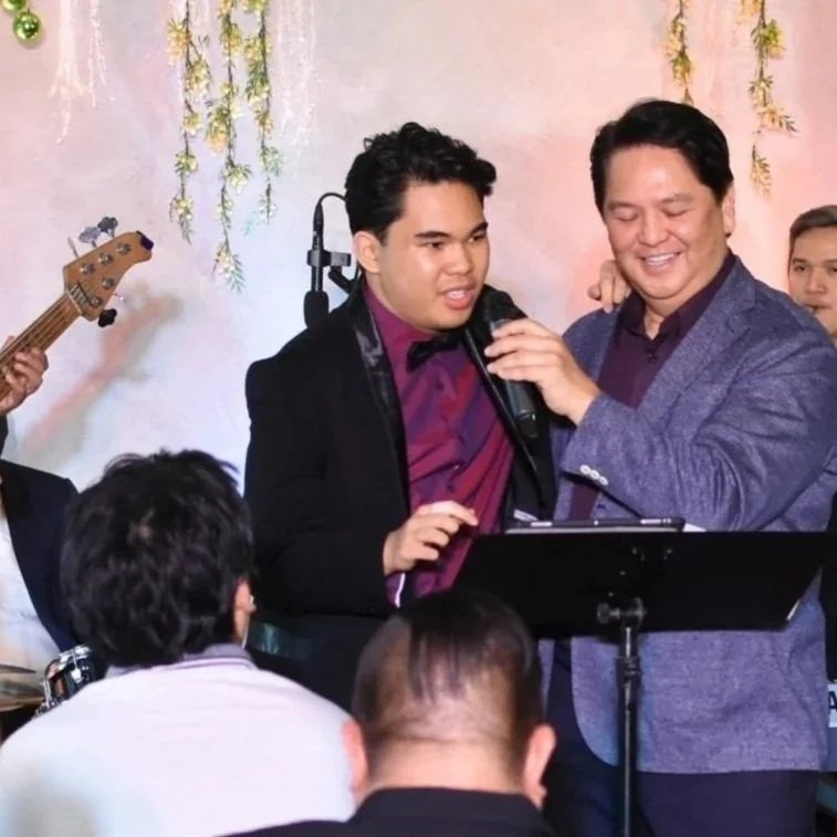 Two men are on stage, singing into a microphone and a community holiday music event. There are several people in the audience, and a full band is behind them with festive decorations.