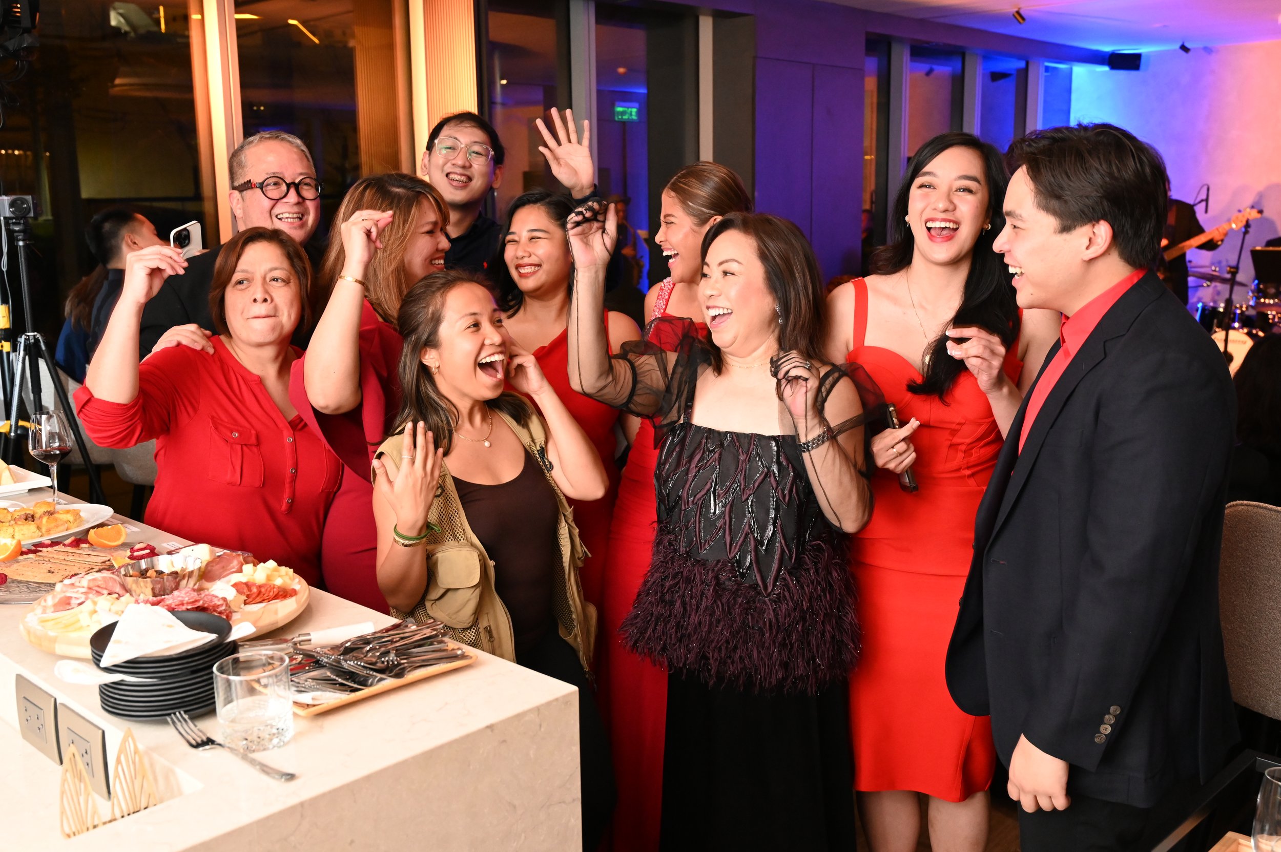 A group of people celebrating at a party, smiling and laughing, with a woman holding a microphone, in a decorated indoor venue with colorful lighting and a food table in the foreground.
