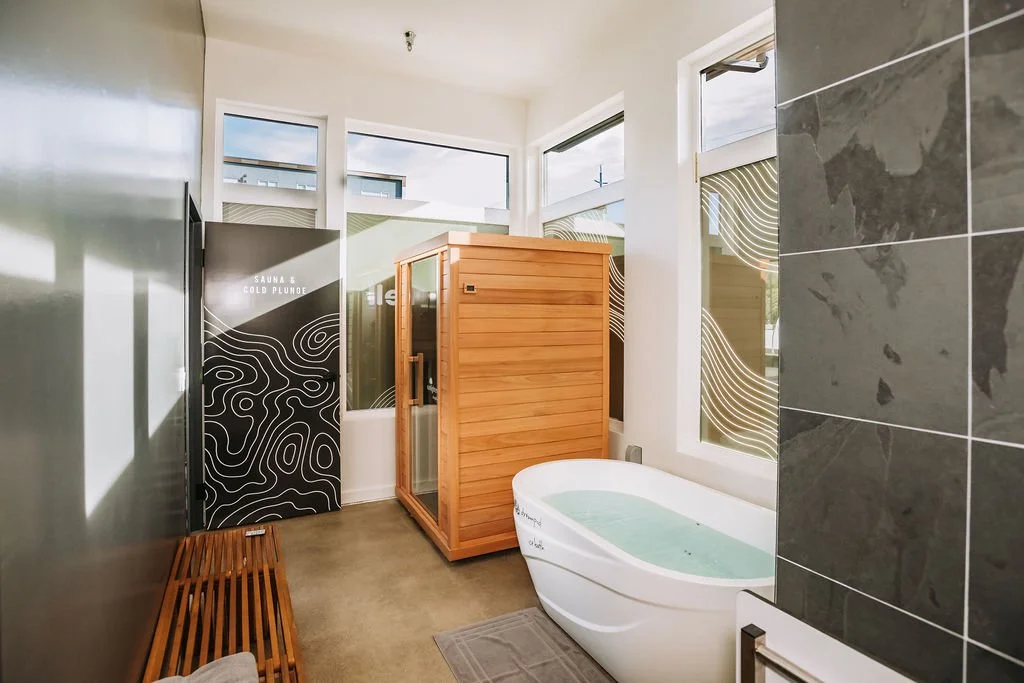 A room with a bathtub filled with water, a wooden sauna cabinet, a black sign with white wavy lines labeled 'Sauna & Cold Plunge', a small wooden bench, and large windows letting in natural light.
