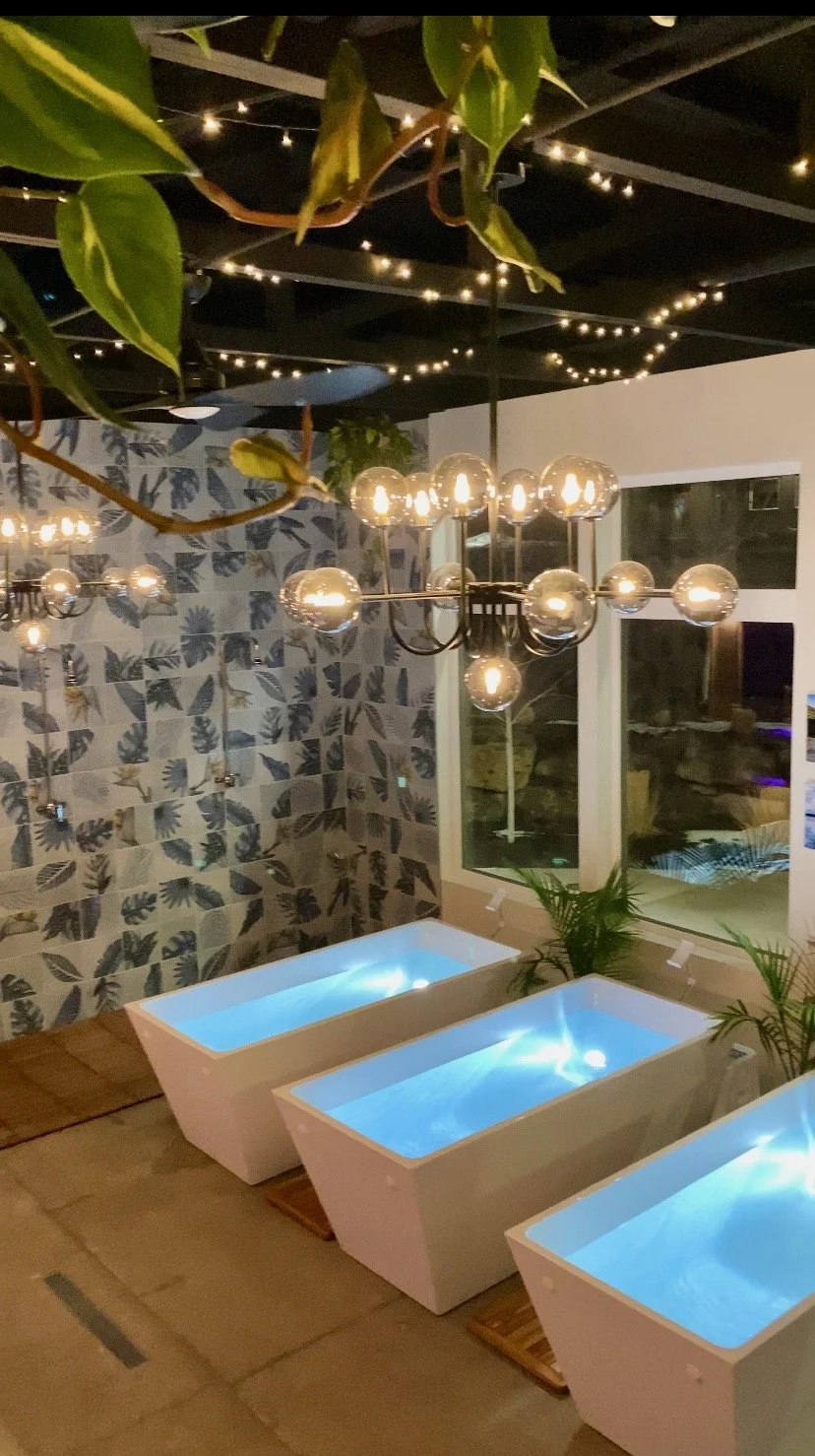 Indoor spa room with three white soaking tubs filled with water illuminated with blue light, decorated with plants, modern light fixture, and ceiling string lights