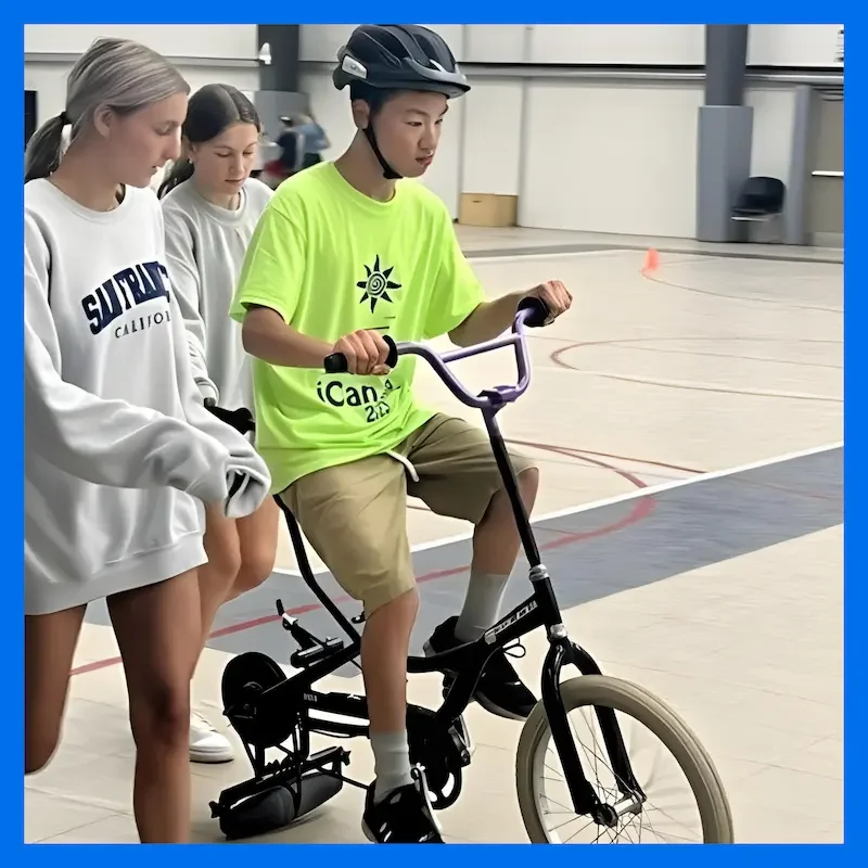 Teen participant in a bright green iCan Bike shirt practices riding with support from volunteers on an indoor track, highlighting adaptive biking and youth sports programming for children with disabilities