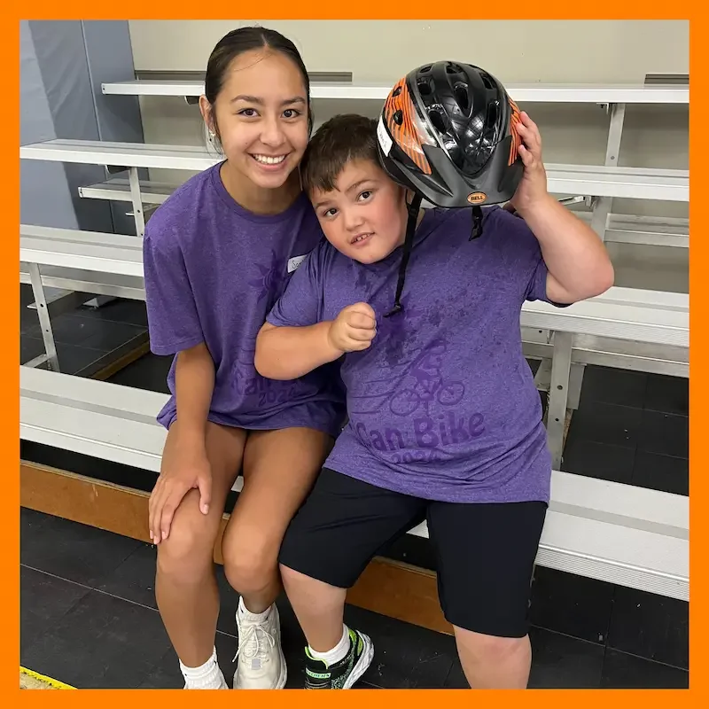 Smiling volunteer and child participant sitting on gym bleachers during a learn to bike program wearing matching “iCan Bike” purple t-shirts. The child proudly holds a bike helmet, highlighting TCP’s youth sports initiatives in Western NY
