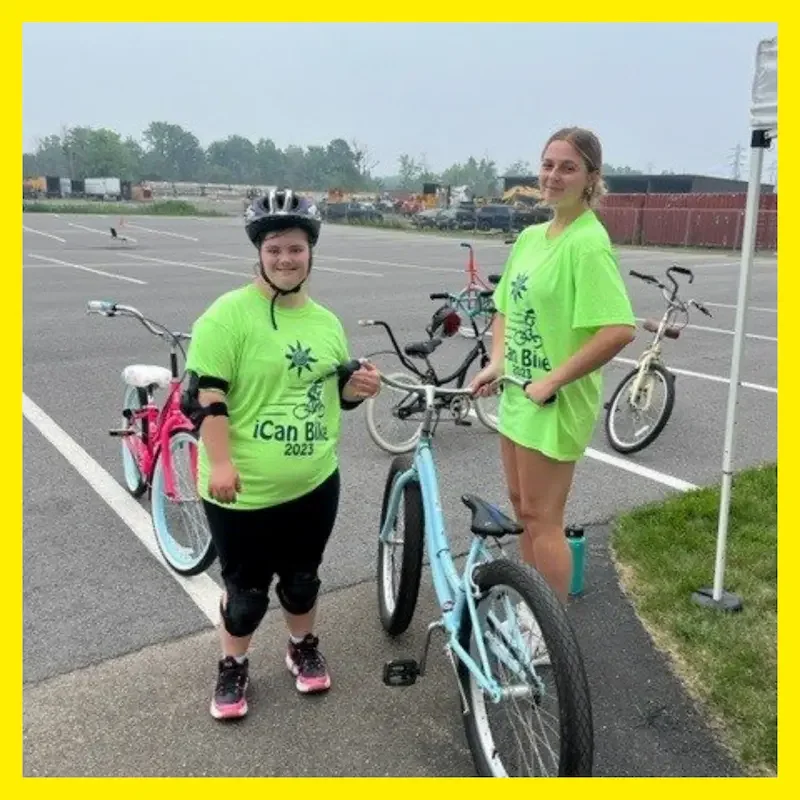 Smiling iCan Bike participant and volunteer in bright green shirts stand beside bicycles in an outdoor lot. The Champion Project ICan Shine Learn to ride a bike camp