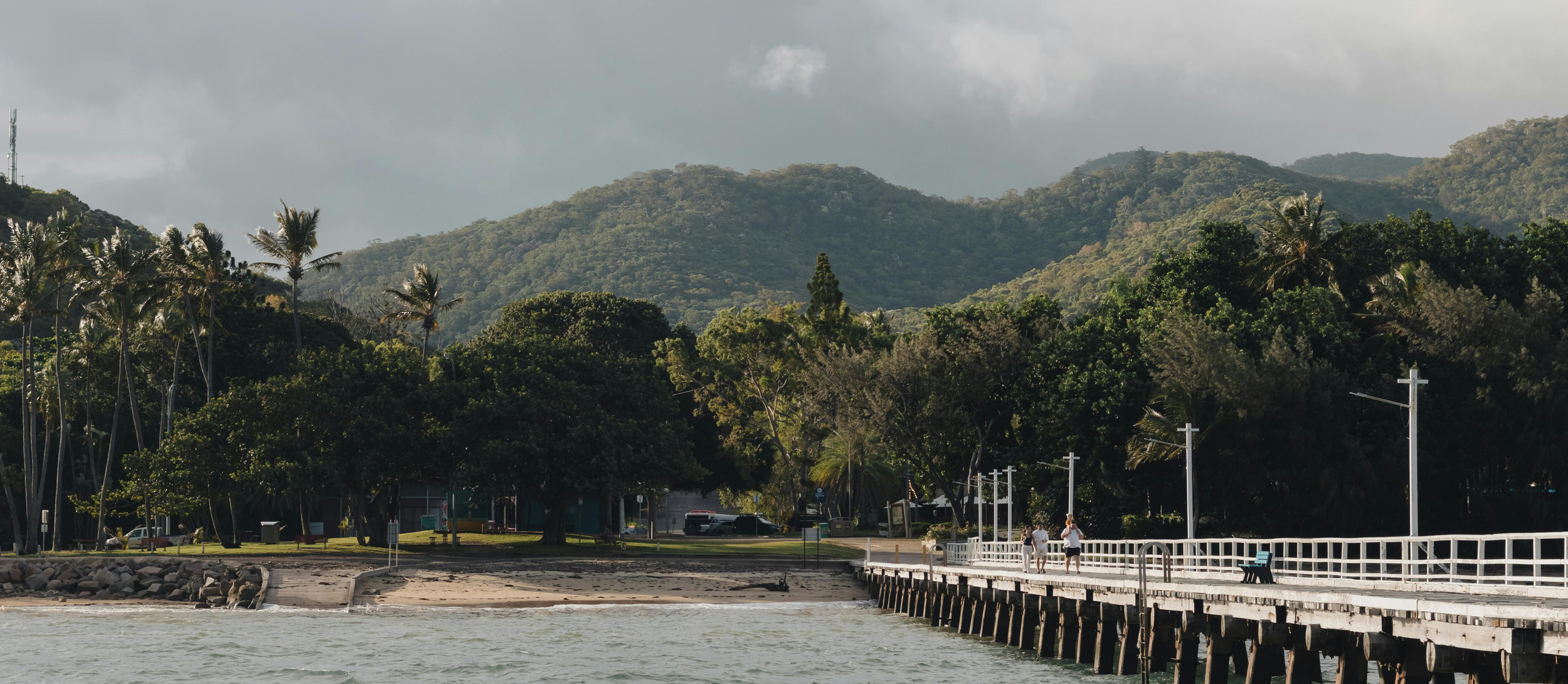 People walking on a pier by the water with lush green trees and hills in the background.