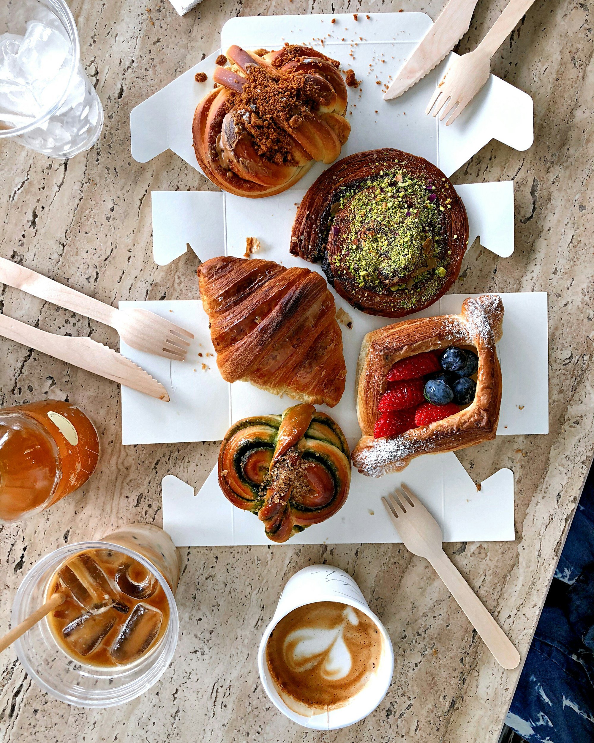 Assorted pastries including a cinnamon roll, croissant, fruit Danish, and a pistachio pastry, along with iced coffee and iced tea on a marble table.
