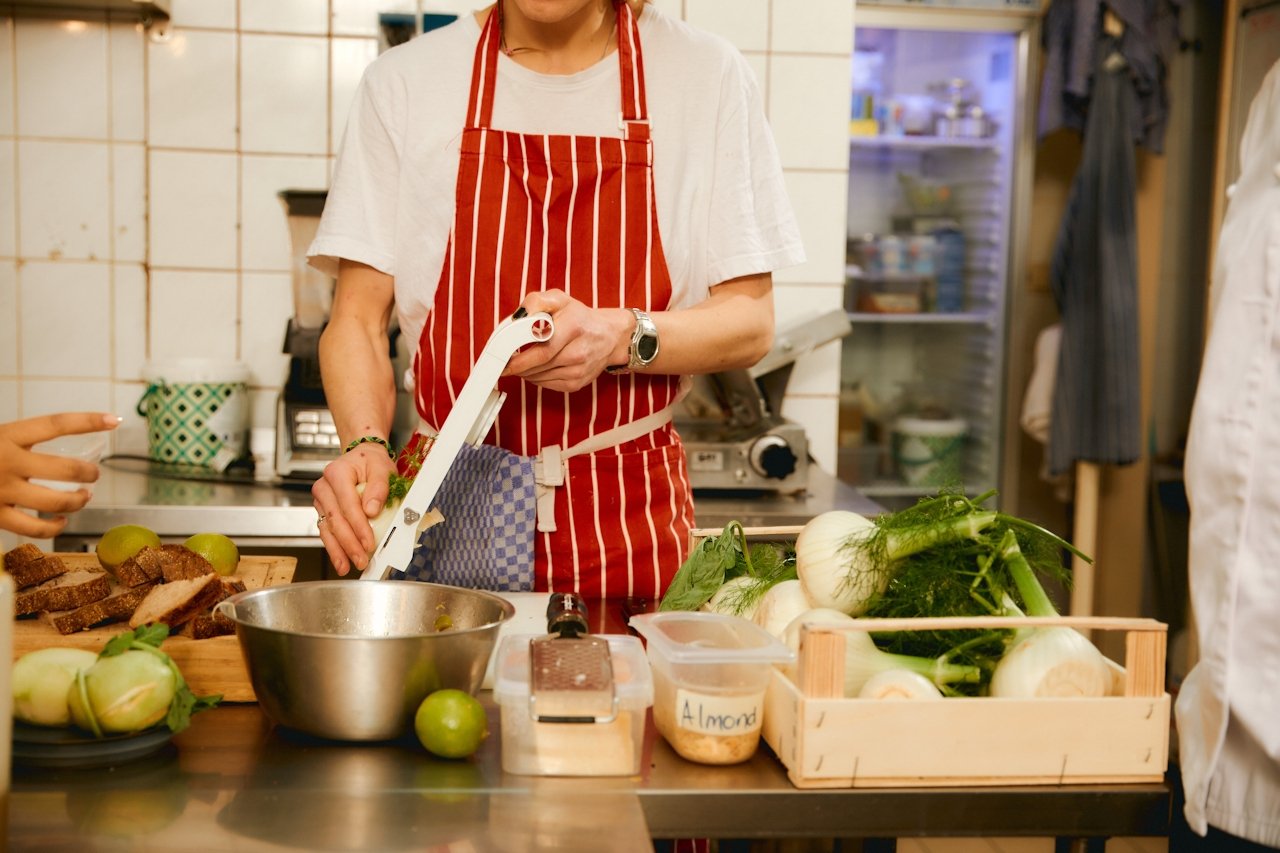 Person wearing a red and white striped apron preparing food in a kitchen, surrounded by onions, limes, and a wooden box labeled 'Almond' on a stainless steel counter.