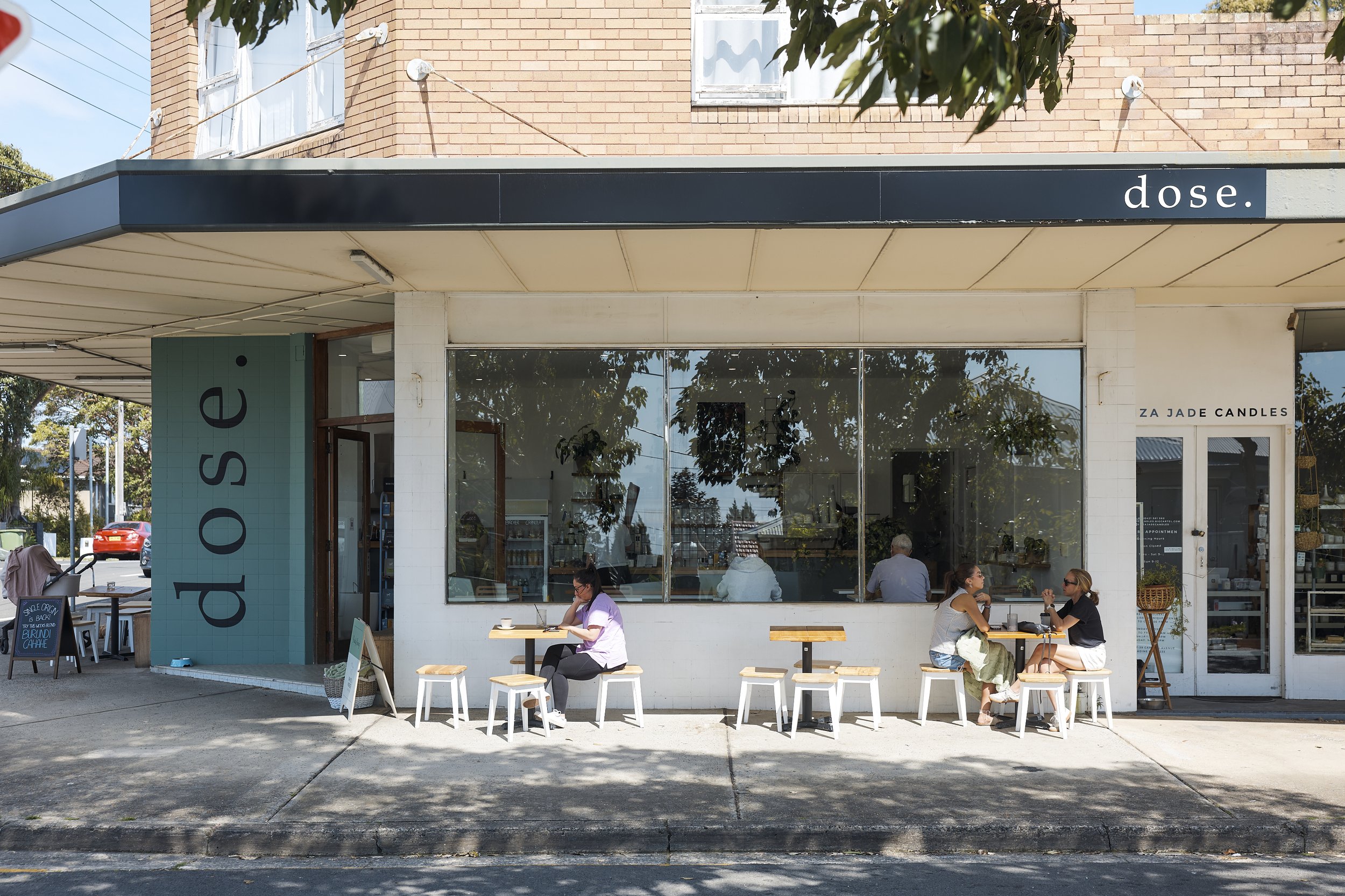 Exterior of a modern café named 'dose' with outdoor seating, three women sitting at a table on the sidewalk, large window reflecting trees, and a brick building above.
