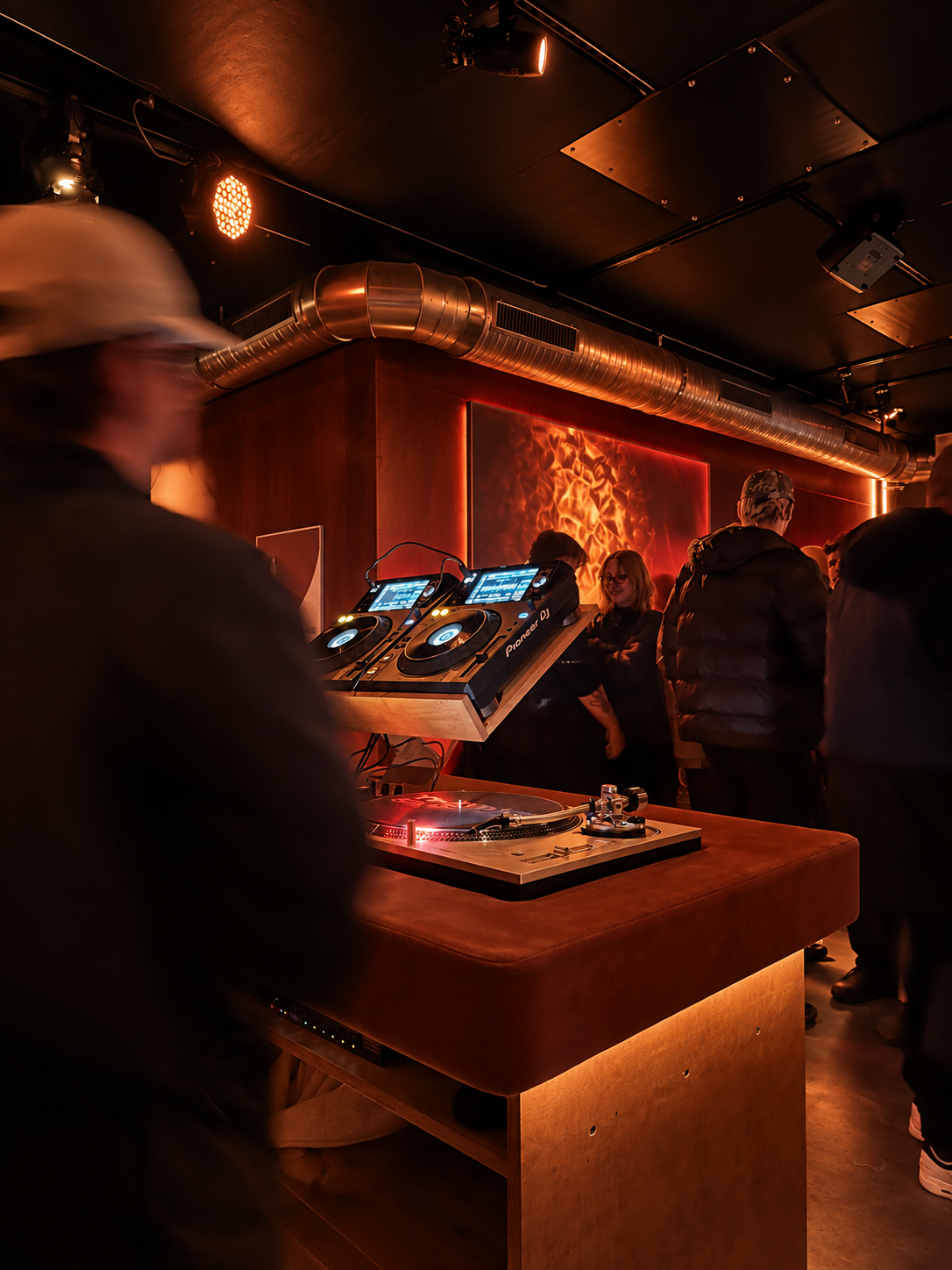 DJ-Equipment auf einem Tanzflächen in einer dunklen Bar oder Club. Menschen stehen im Hintergrund, eine Wand mit rotem Feuerbild im Hintergrund.