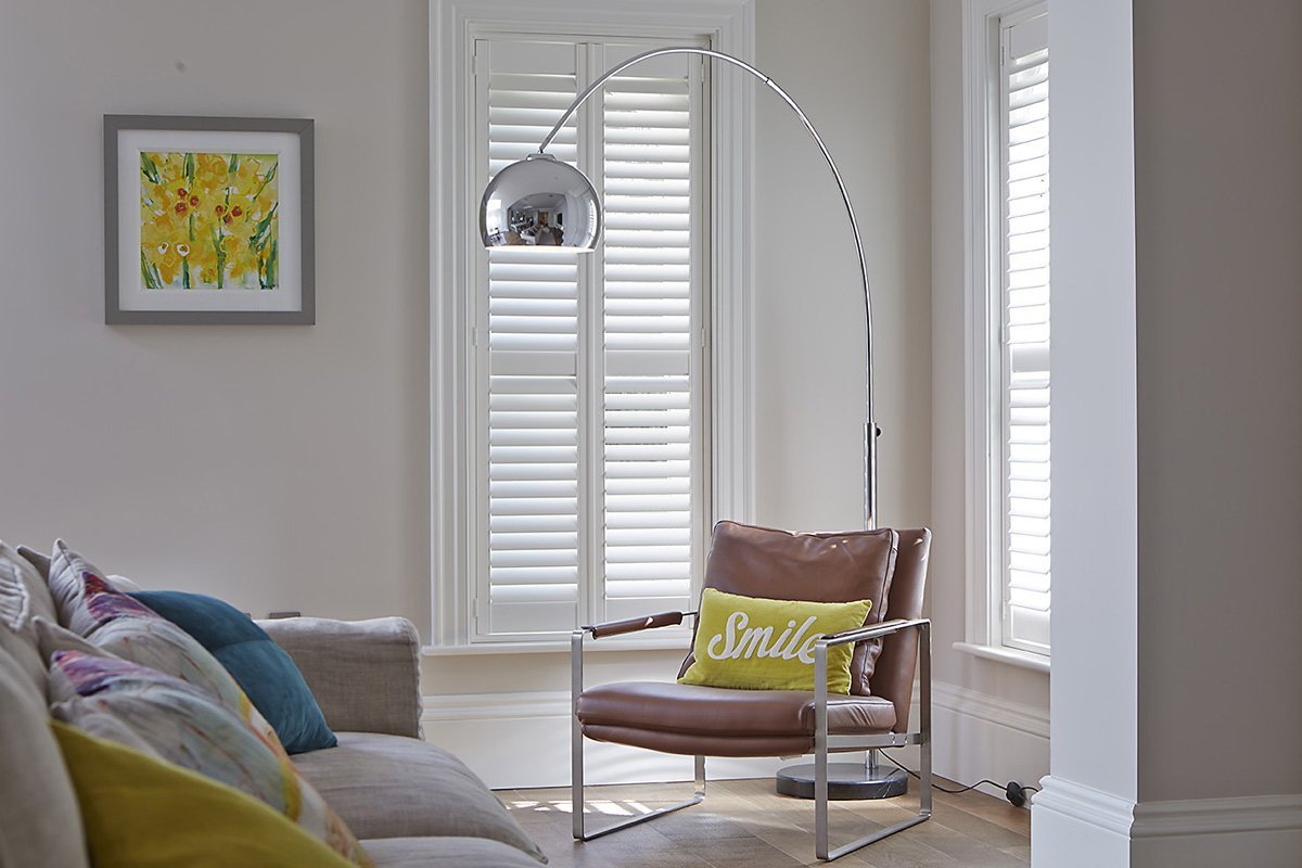 A living room with white walls, wooden floors, and large windows with white shutters. There is a gray sofa with colorful pillows, a brown leather armchair with a yellow pillow that says "Smile," and a tall chrome floor lamp with a dome-shaped shade.