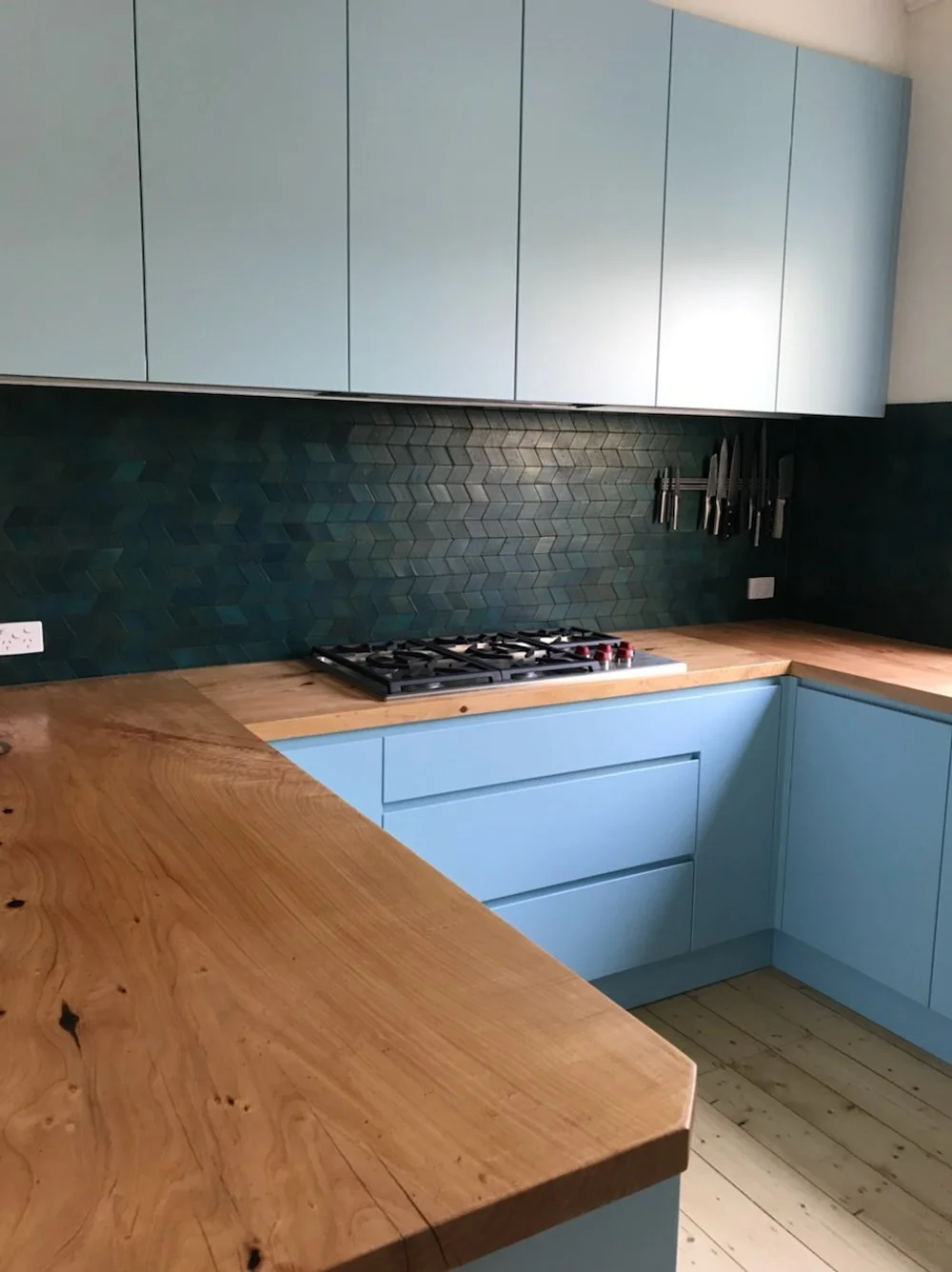 View of the kitchen countertop, showing wood finish bench tops, green tiles, blue cabinets. Modern and functional. Works well with natural light.