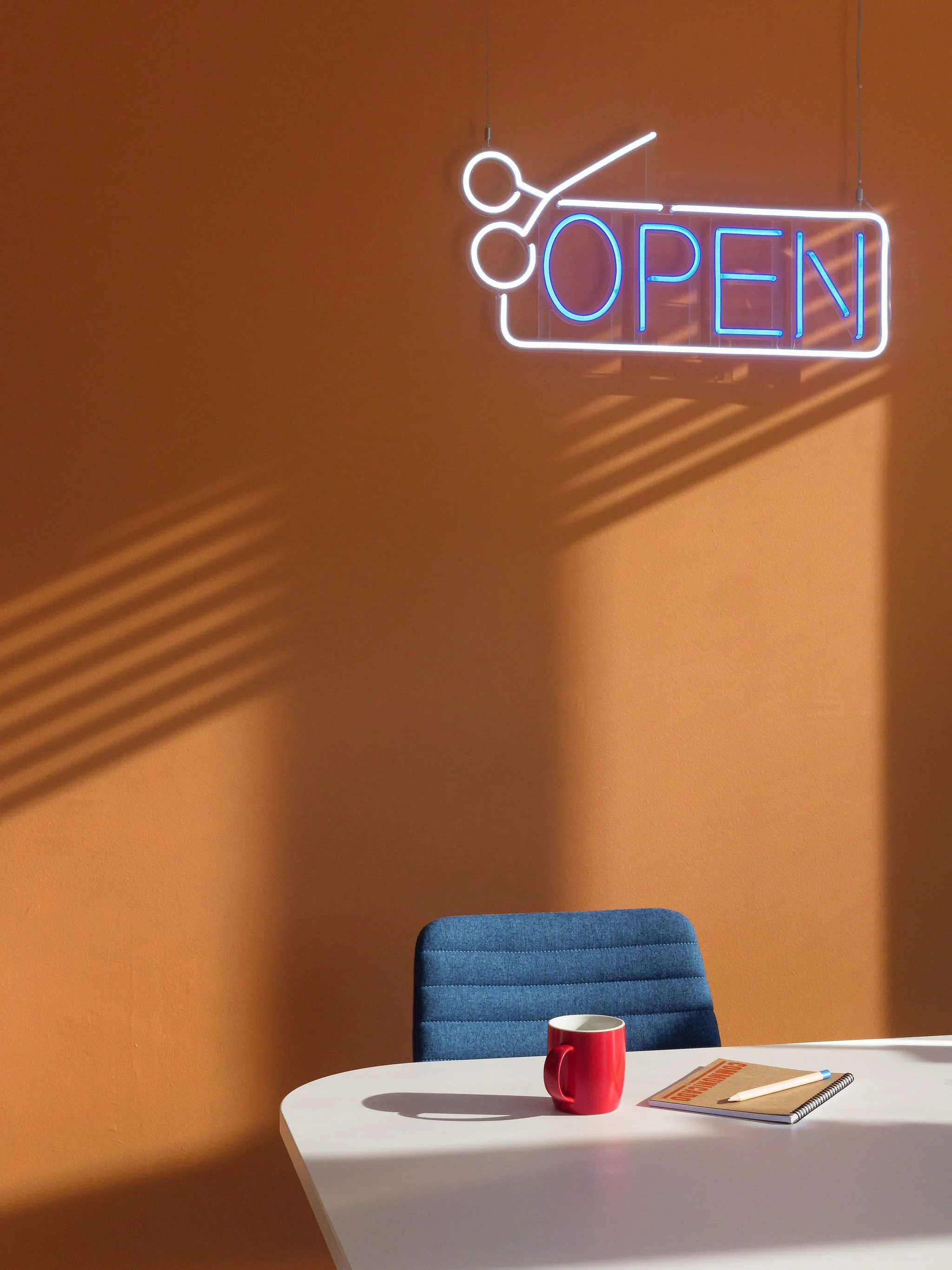 Tall photo of an office interior, showing a white desk with blue chair, behind is an orange wall decorated with a bright neon OPEN sign